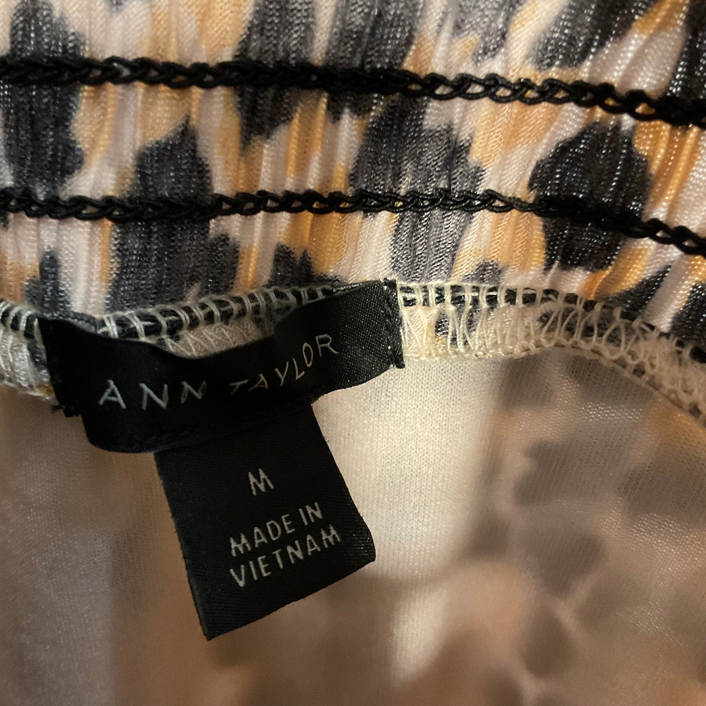 Close-up of a garment's inner neck area showing a black brand tag that reads 'ANN ZAYLOR' and 'M MADE IN VIETNAM' over a beige, black, and gray patterned fabric.
