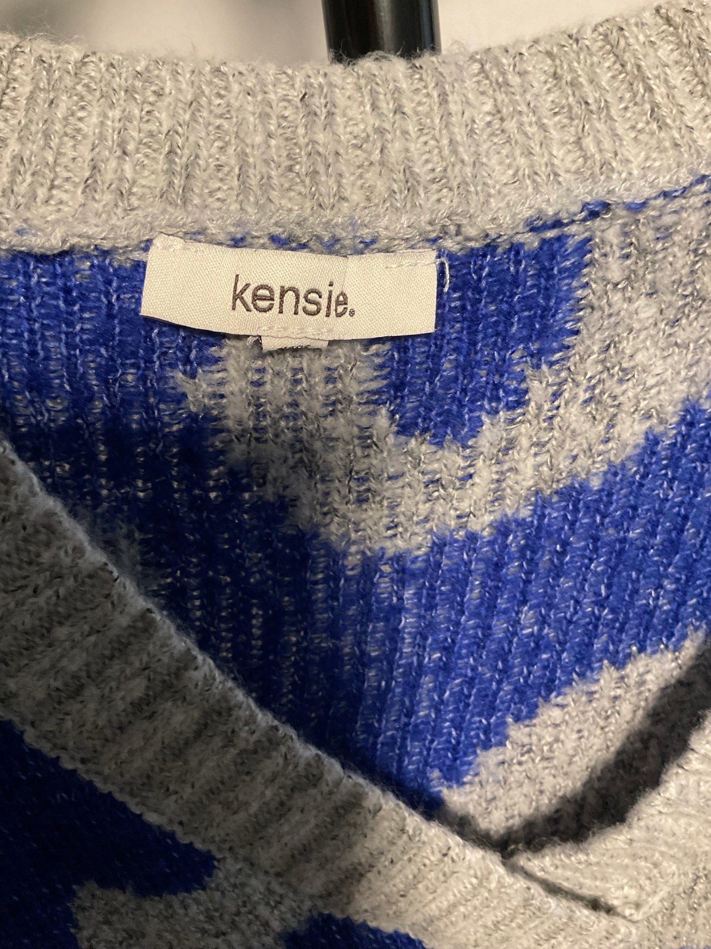 Close-up of a knit sweater showing a cream brand label that reads 'kensie.' The garment has a blue and heather gray patterned knit design.