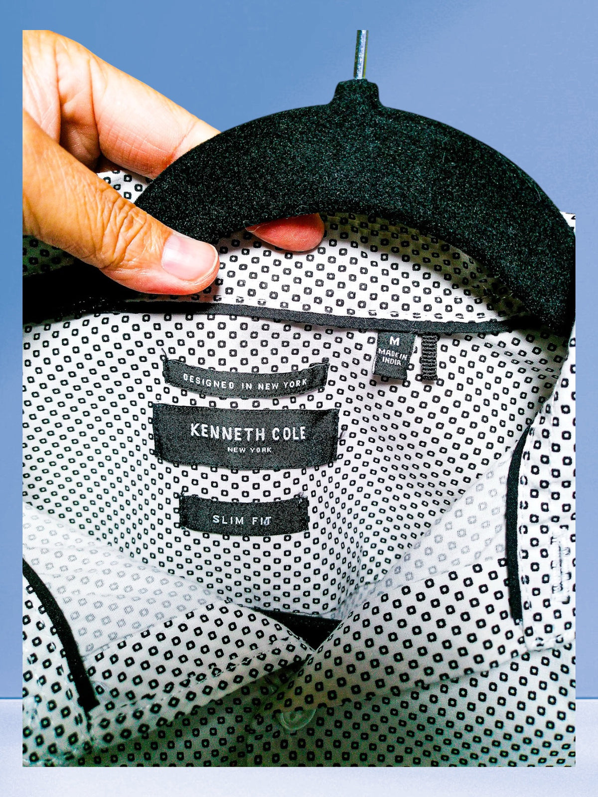 A close-up of a Kenneth Cole Slim Fit dress shirt with a black and white micro-dot pattern, held on a black hanger by a hand. The brand label is clearly visible inside the collar.