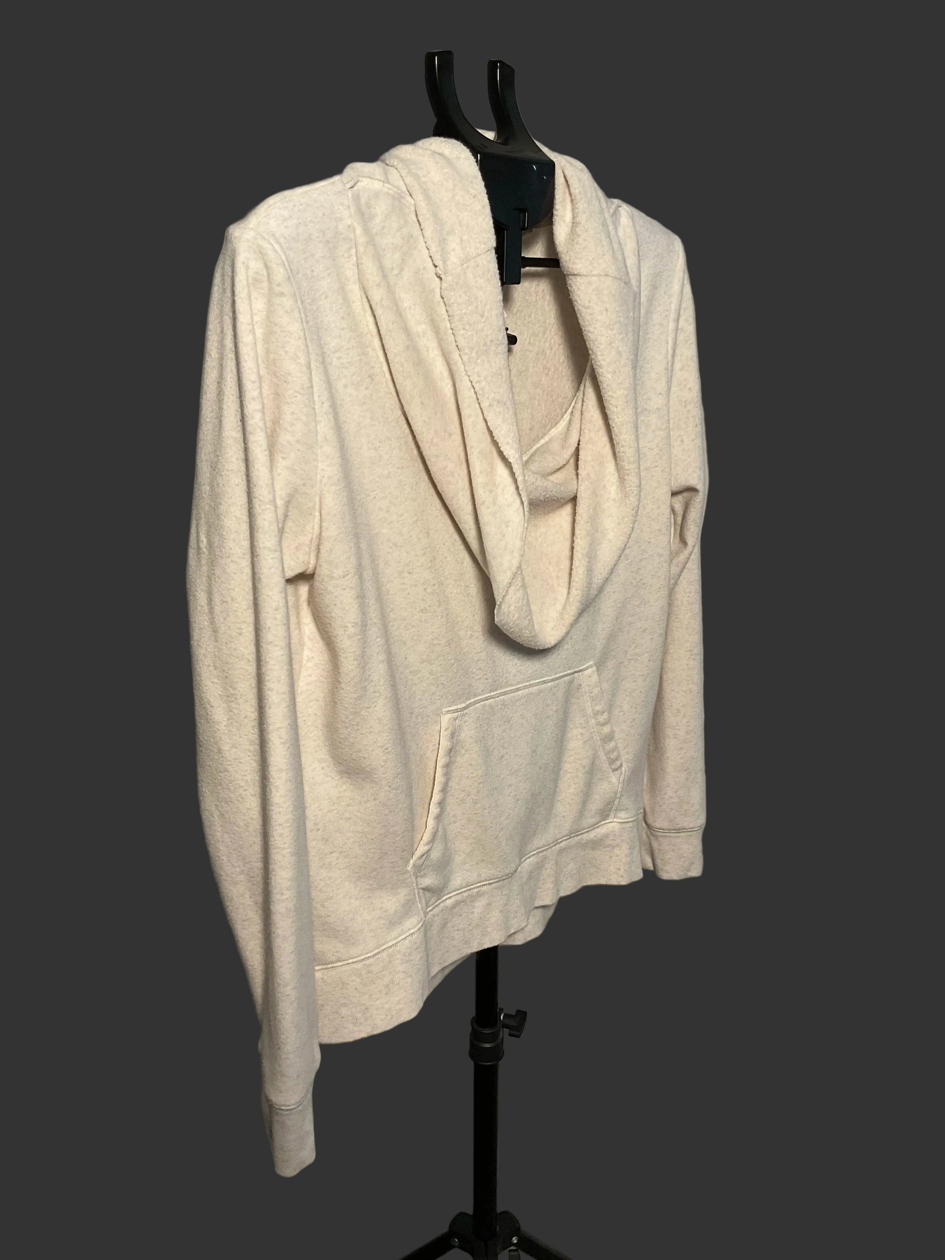 A cream-colored cowl neck pullover hoodie with a front kangaroo pocket, displayed on a black hanger stand against a plain gray wall.