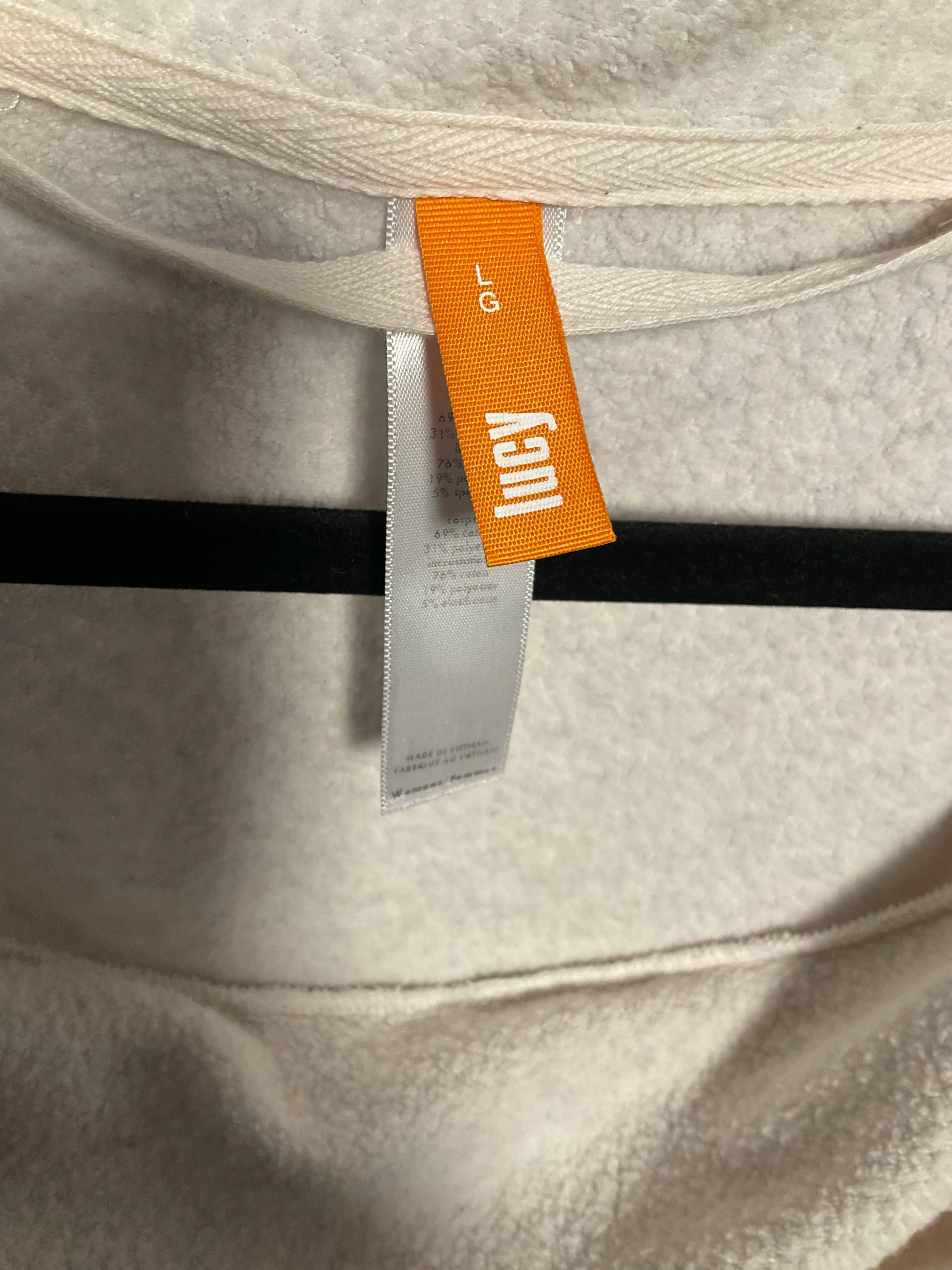 Close-up of a light beige fleece or sherpa garment with a visible orange and white 'lucy' brand tag and a white size tag, hanging against a black hanger.