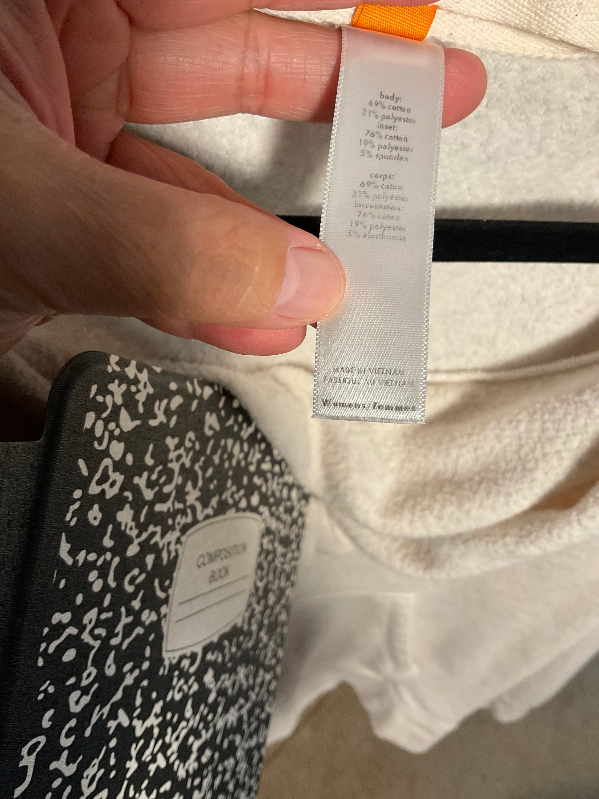 A close-up of a hand holding a white garment tag with printed composition details, positioned over a cream-colored textured fabric. A black and white patterned notebook is visible in the lower-left corner.