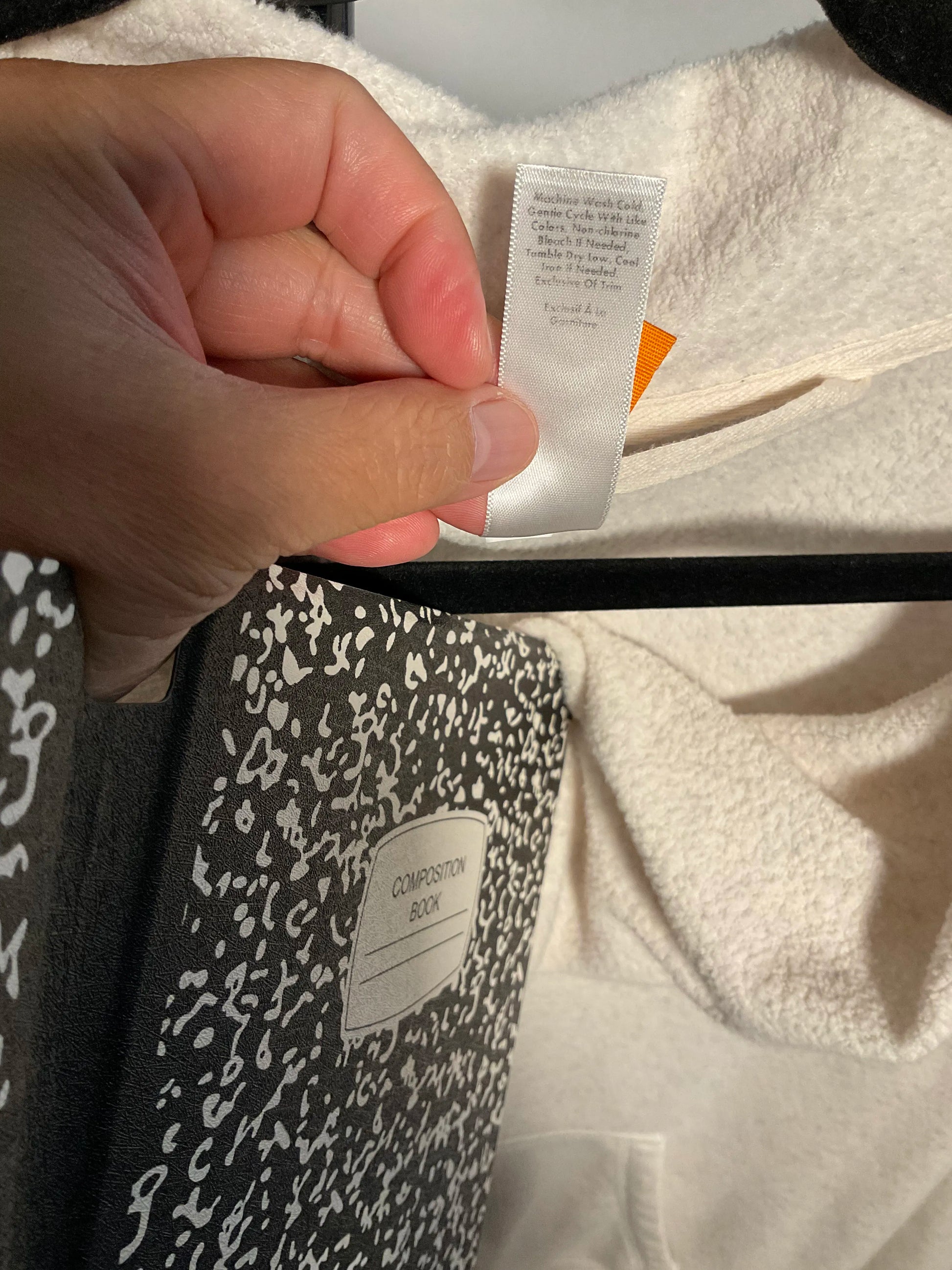 A close-up of a hand holding a white fabric care label inside a cream-colored garment, with a black garment featuring a white speckled pattern partially visible below.