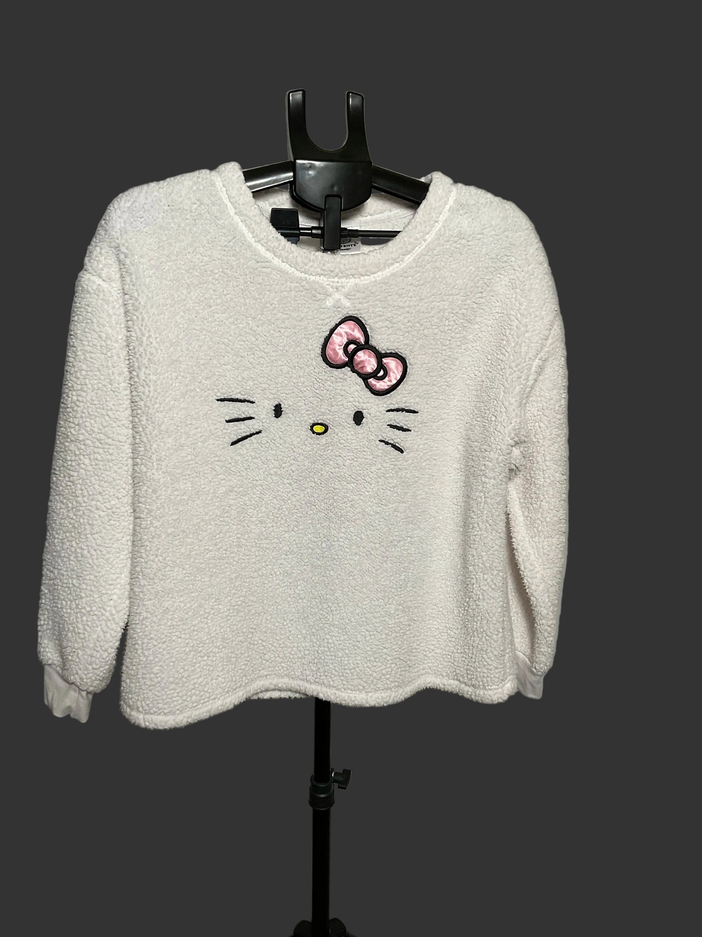 A white fuzzy long-sleeve sweatshirt featuring an embroidered Hello Kitty face and bow on the front, hanging on a black hanger against a gray studio backdrop.