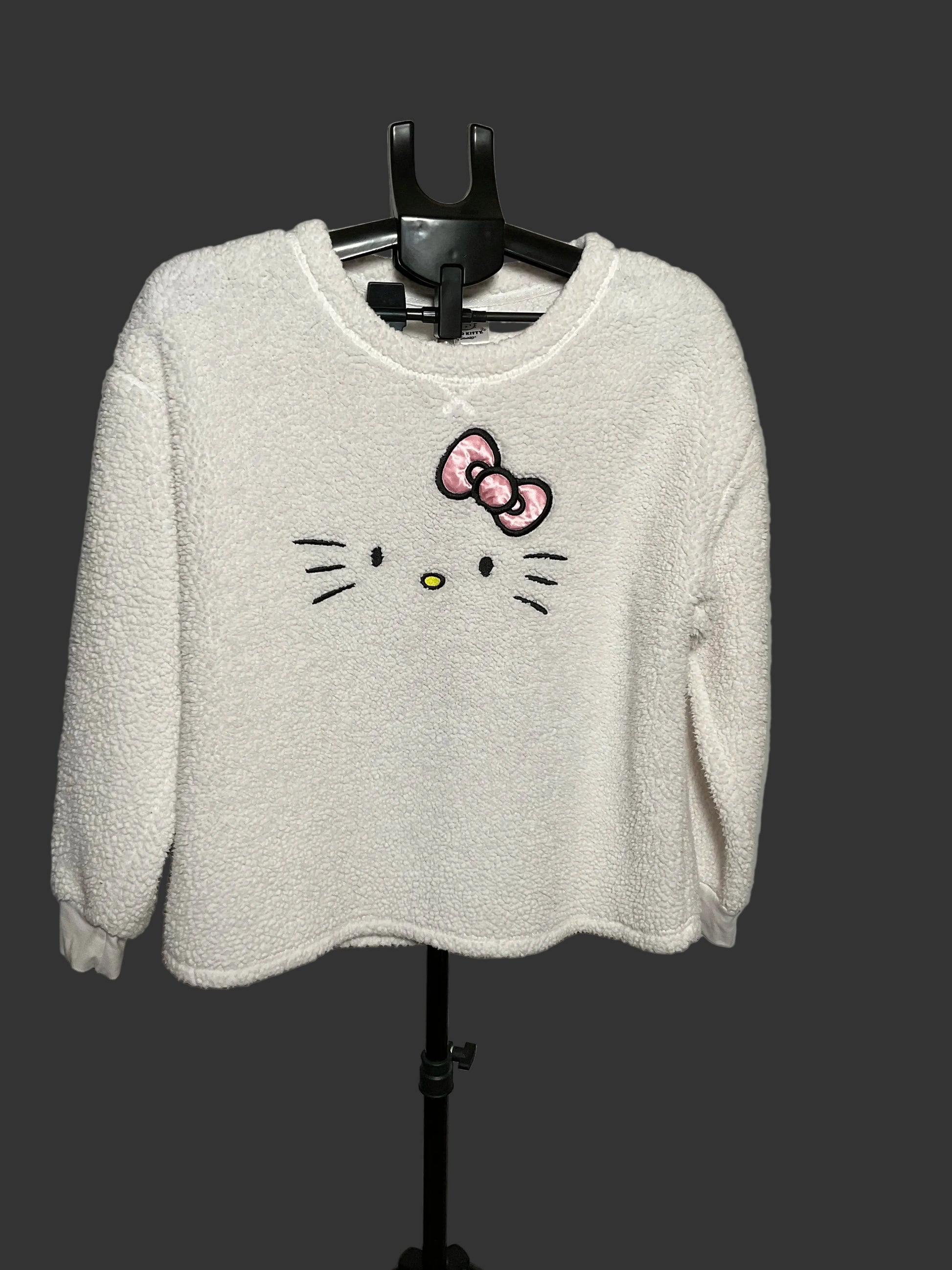 A white fuzzy long-sleeve sweatshirt featuring an embroidered Hello Kitty face and bow on the front, hanging on a black hanger against a gray studio backdrop.