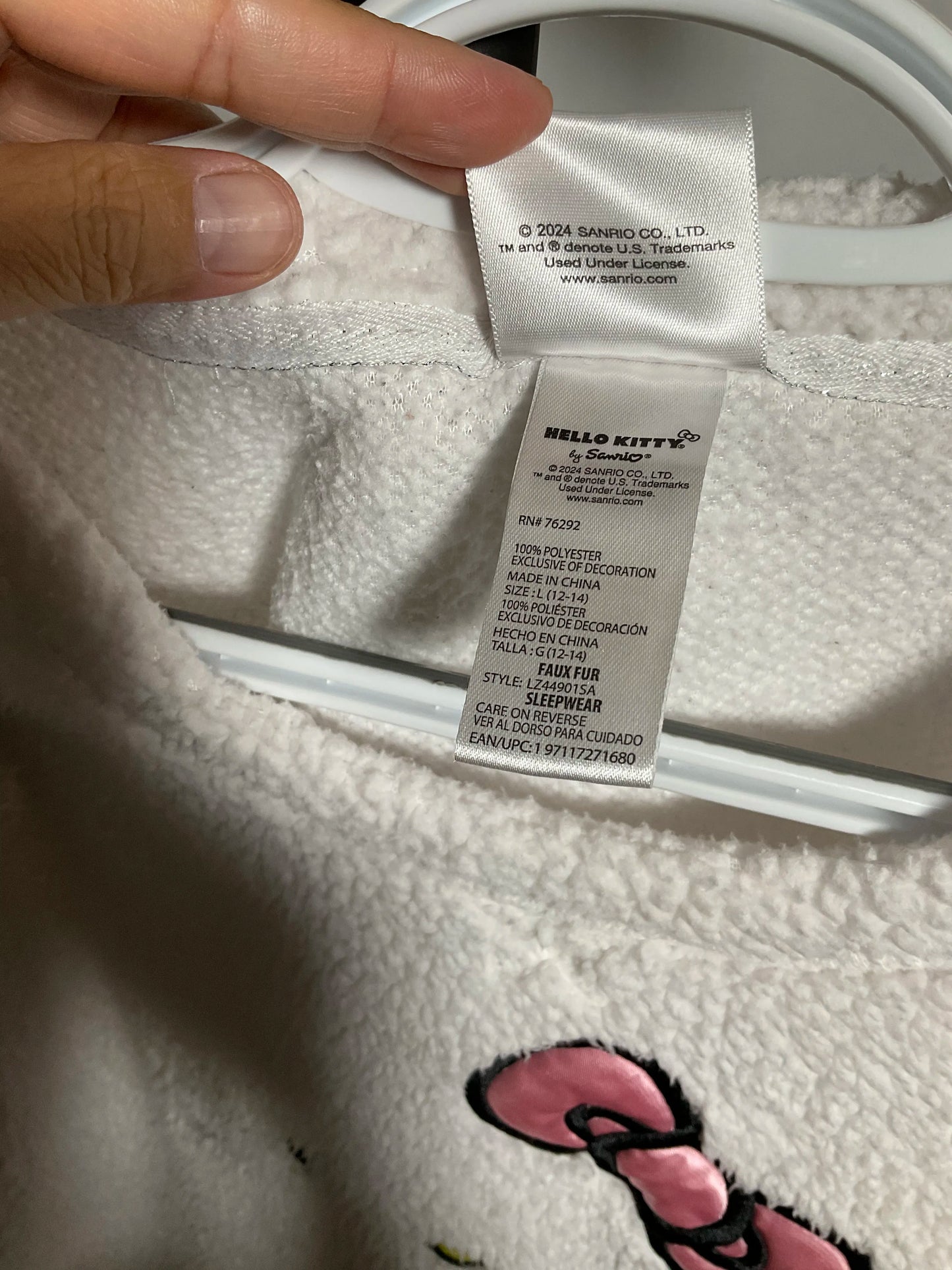 Close-up of a white plush Hello Kitty garment with a visible interior care label and a hand holding the collar area. A pink Hello Kitty bow graphic is visible on the fabric.