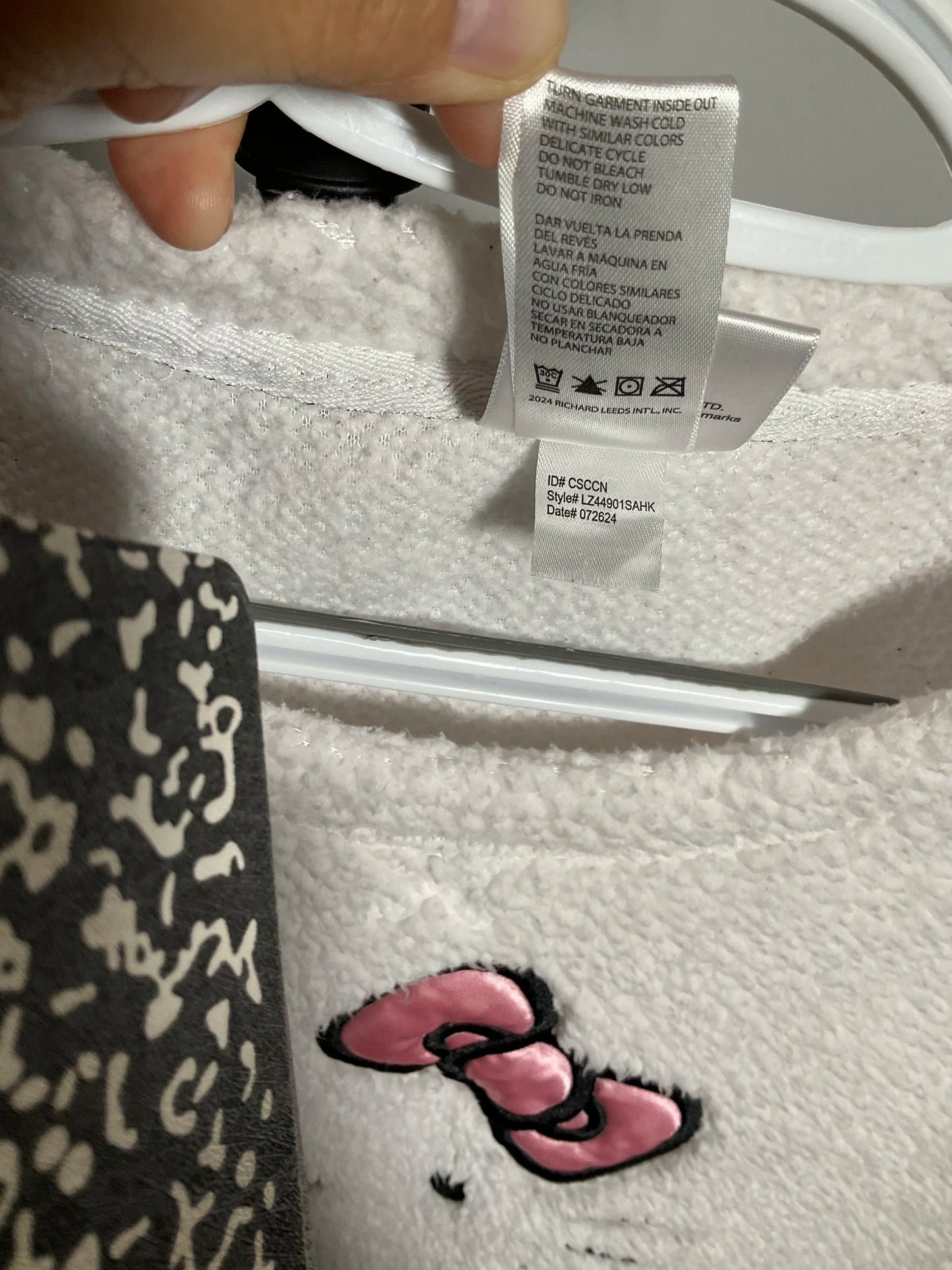 Close-up of a white plush fleece garment with a pink bow embroidered near the hem, showing a sewn-in care label and part of a black-and-white patterned garment beside it.