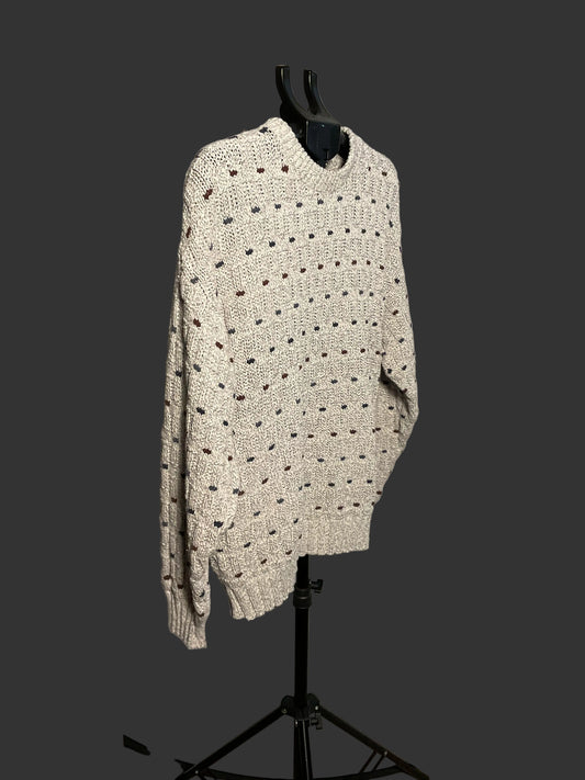 A cream-colored chunky knit sweater with dark brown dotted details, displayed on a black hanger stand against a white circular studio backdrop.