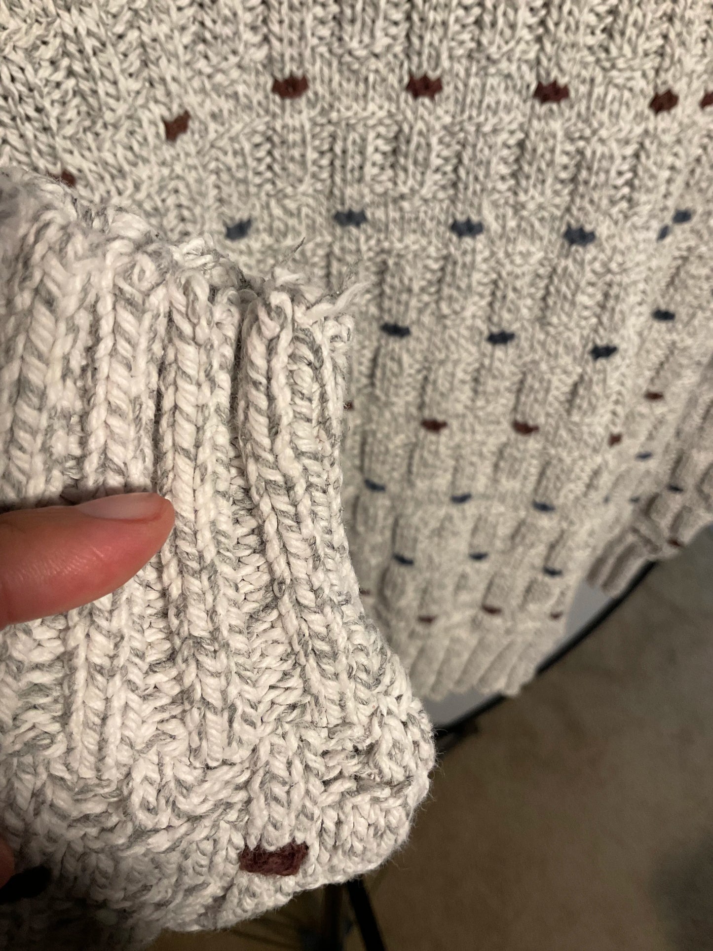 Close-up of a hand holding up a ribbed knit sleeve of a chunky knit sweater, showing the texture and color of the cream-colored yarn with small scattered brown and blue accents.