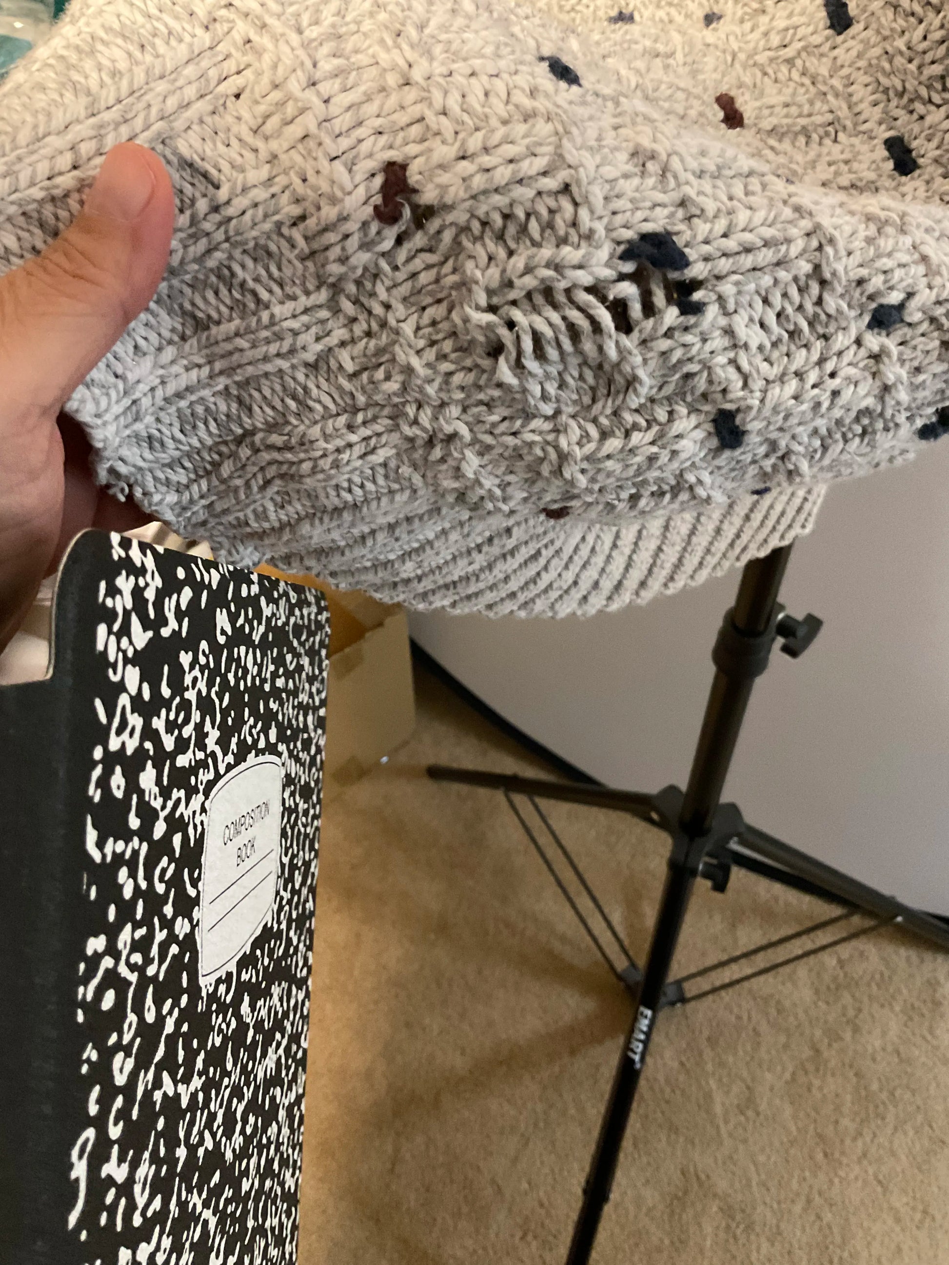 A close-up of a hand holding up a chunky knit blanket or throw in a light gray color with small dark and reddish-brown flecks. A black-and-white speckled notebook is visible in the lower-left corner, and a studio light stand is in the background.