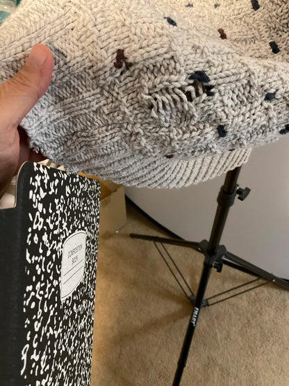 A close-up of a hand holding up a chunky knit blanket or throw in a light gray color with small dark and reddish-brown flecks. A black-and-white speckled notebook is visible in the lower-left corner, and a studio light stand is in the background.