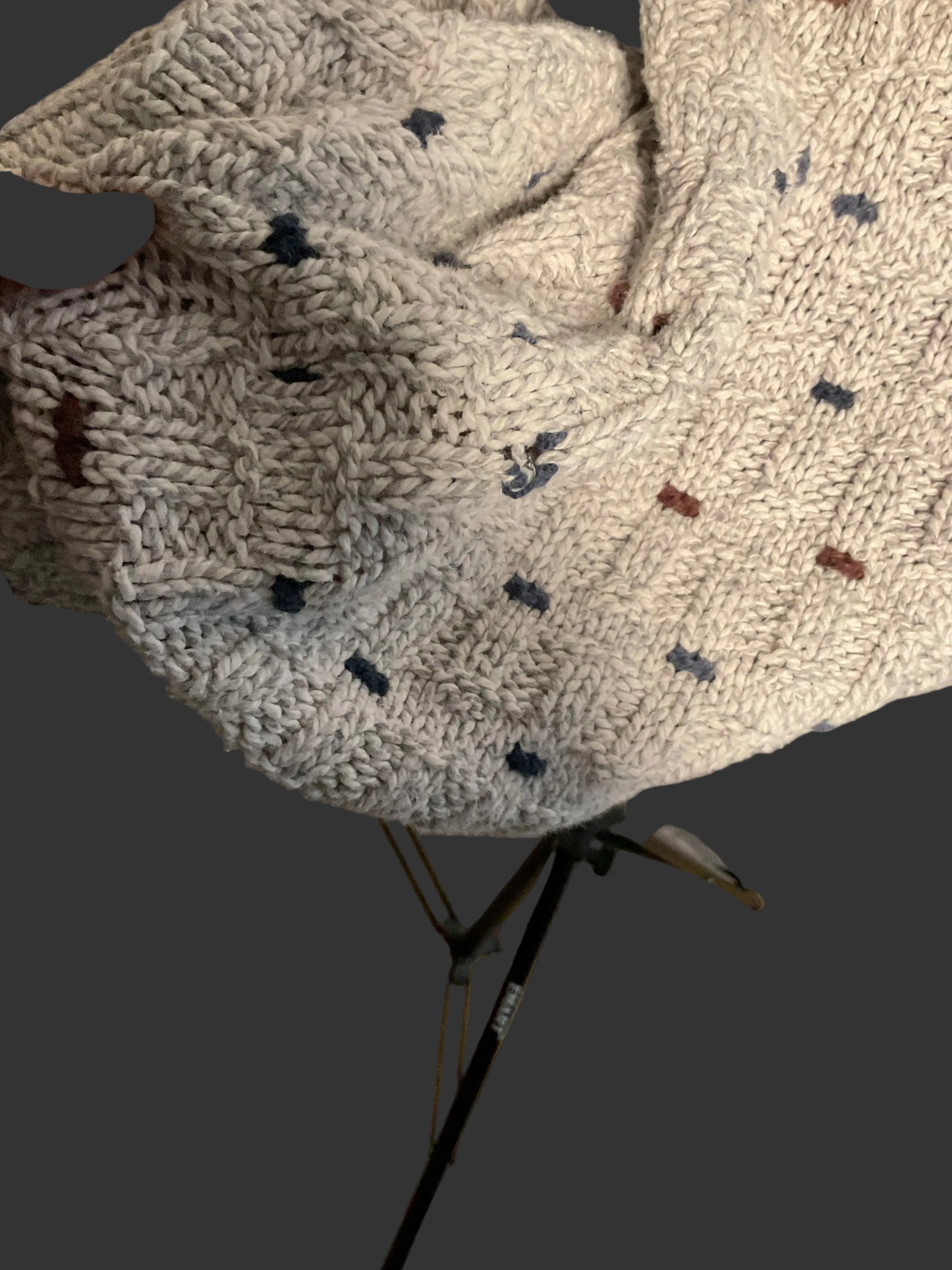 A close-up of a chunky knit sweater or hat in a heathered off-white/cream yarn with small scattered dark navy and rust-brown accents, held by a hand. A JBL studio light stand is visible in the background.