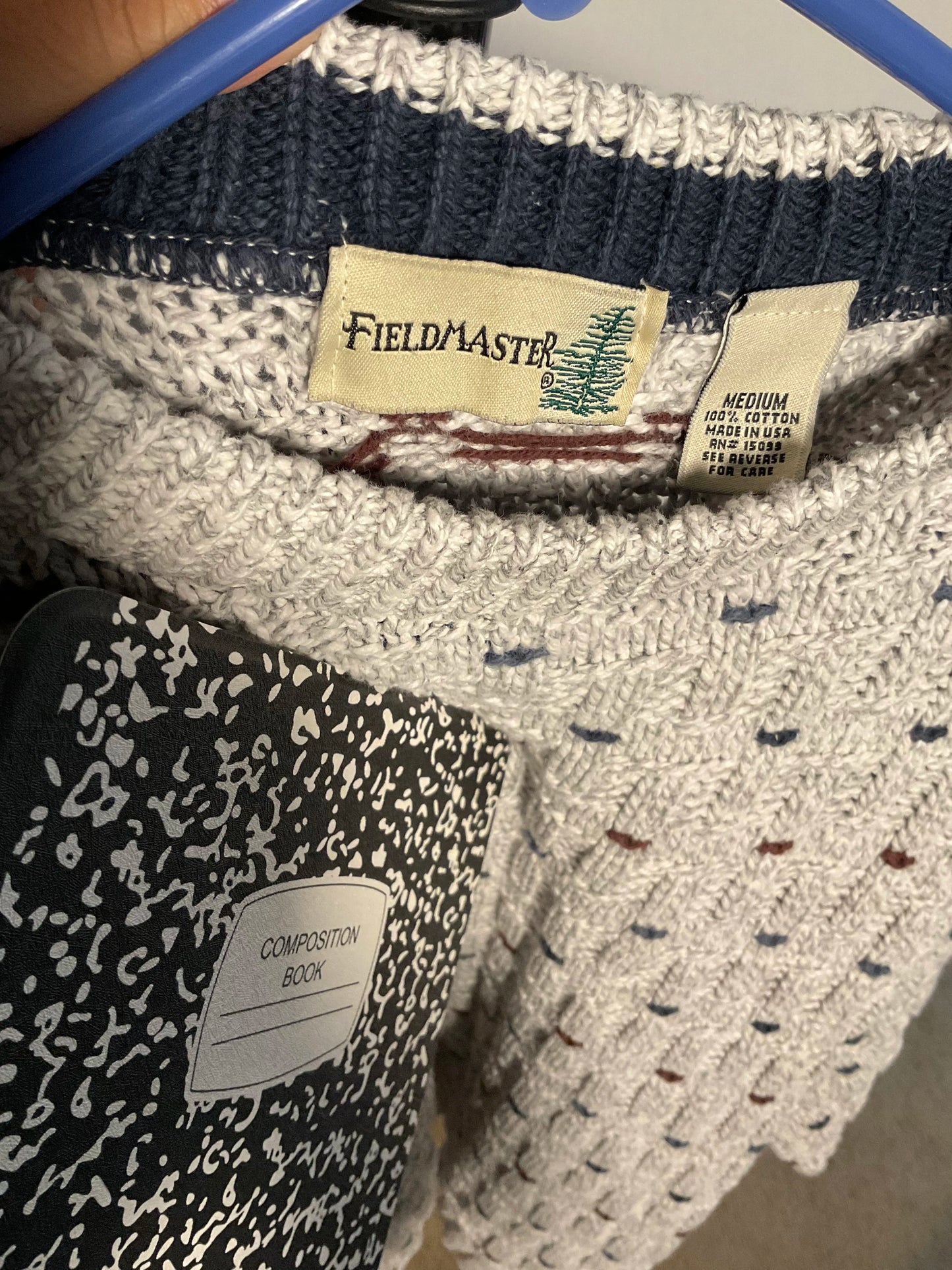 Close-up of a knit sweater inside a blue hanger, showing a cream and navy ribbed collar with a visible Fieldmaster neck label. A black-and-white patterned composition book is partially covering the lower left area.