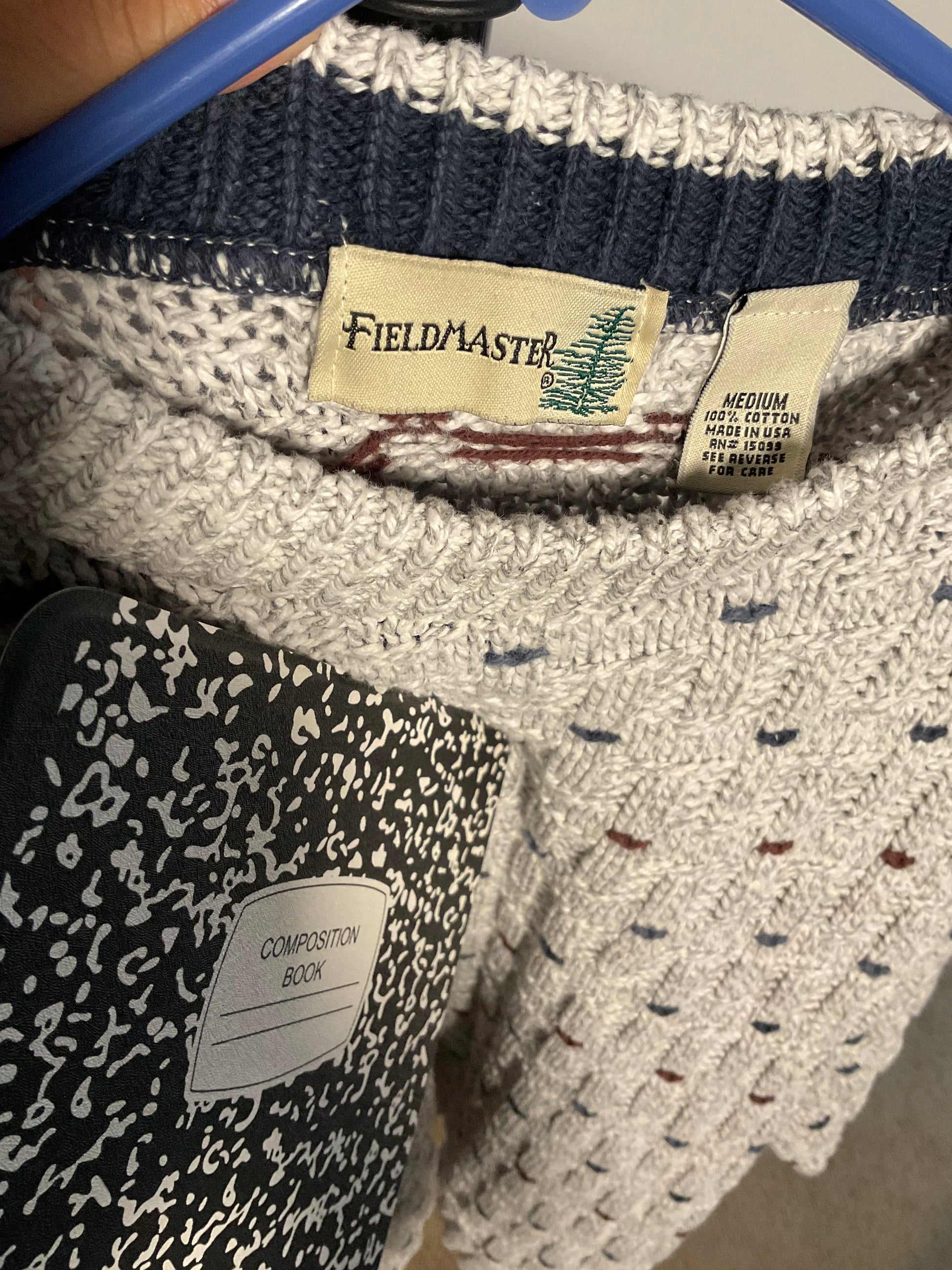 Close-up of a knit sweater inside a blue hanger, showing a cream and navy ribbed collar with a visible Fieldmaster neck label. A black-and-white patterned composition book is partially covering the lower left area.