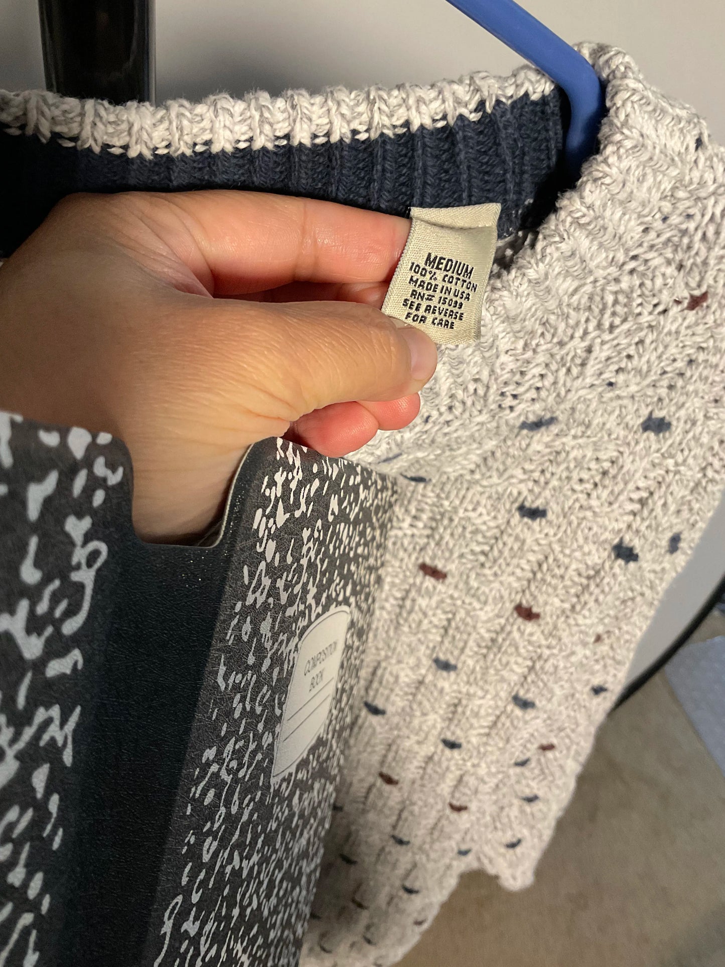 A close-up of a person's hand holding a size 'MEDIUM' fabric tag on a knit sweater. The sweater features a heathered light gray and white body with small multicolored flecks, a dark navy ribbed collar, and a dark gray sleeve with a white abstract pattern.