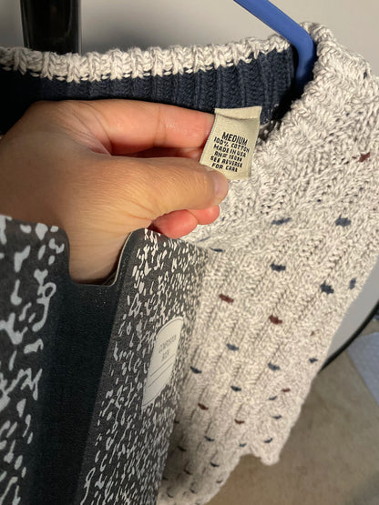 A close-up of a person's hand holding a size 'MEDIUM' fabric tag on a knit sweater. The sweater features a heathered light gray and white body with small multicolored flecks, a dark navy ribbed collar, and a dark gray sleeve with a white abstract pattern.