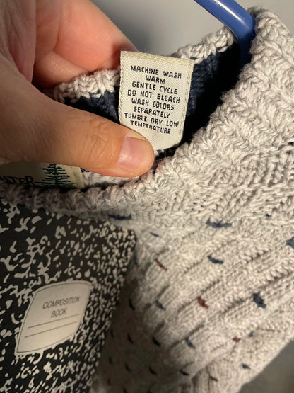 A close-up of a knit sweater's interior neck label held by a hand. The label reads 'MACHINE WASH WARM GENTLE CYCLE DO NOT BLEACH WASH COLORS SEPARATELY TUMBLE DRY LOW TEMPERATURE.