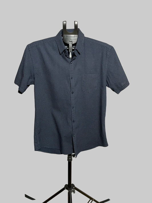 A short-sleeve button-up shirt with a small white geometric pattern on a dark navy base, displayed on a black mannequin stand in front of a white circular studio backdrop.