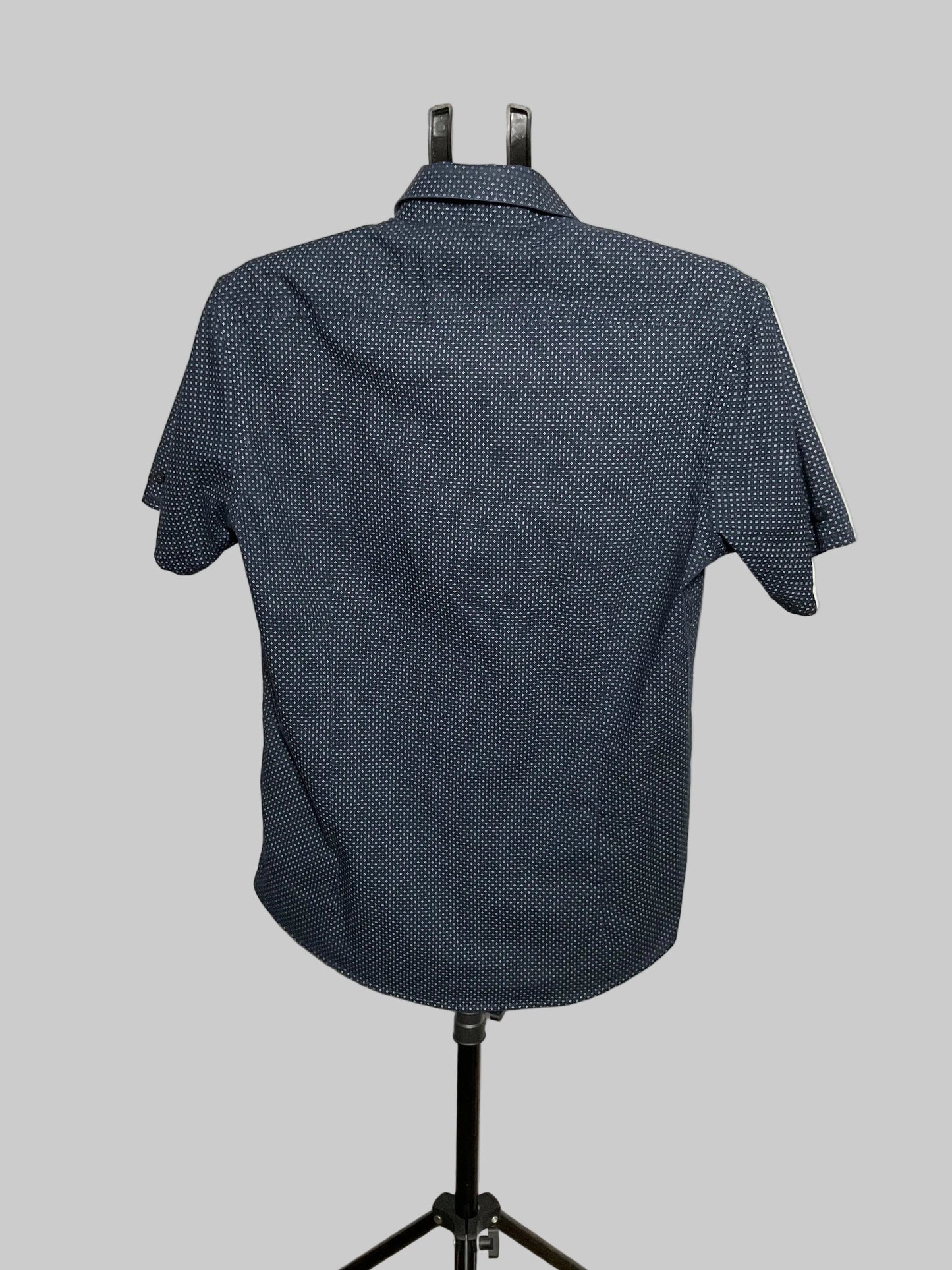 A short-sleeve button-up shirt with a small white geometric pattern on a dark navy base, displayed on a black mannequin stand against a white studio backdrop.