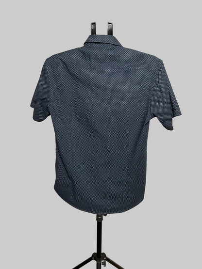 A short-sleeve button-up shirt with a small white geometric pattern on a dark navy base, displayed on a black mannequin stand against a white studio backdrop.