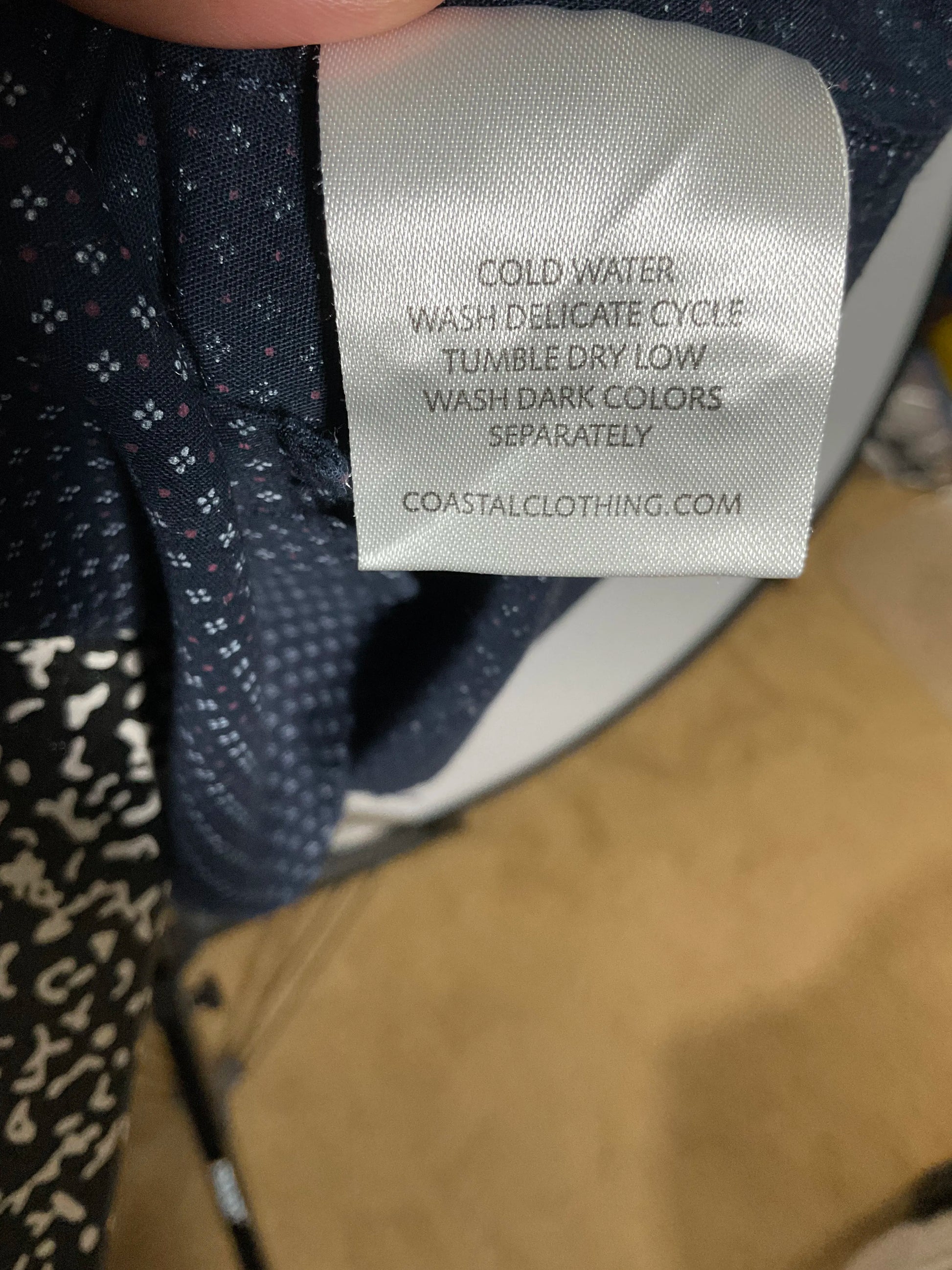 Close-up of a garment's inner neck label reading 'COLD WATER WASH DELICATE CYCLE TUMBLE DRY LOW WASH DARK COLORS SEPARATELY COASTALCLOTHING.COM' attached to dark navy patterned fabric.