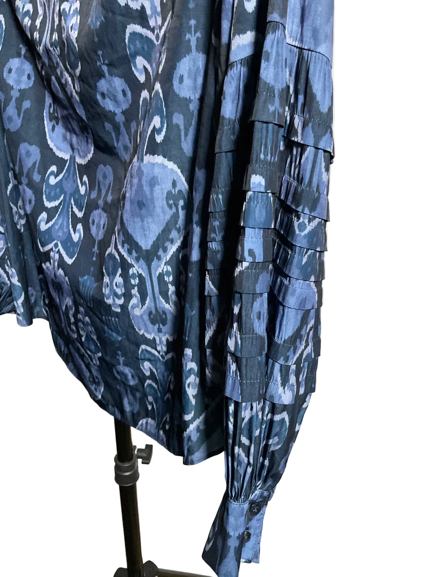 A close-up of a blue patterned blouse with layered ruffle sleeves, displayed on a black dress form stand against a white studio backdrop.