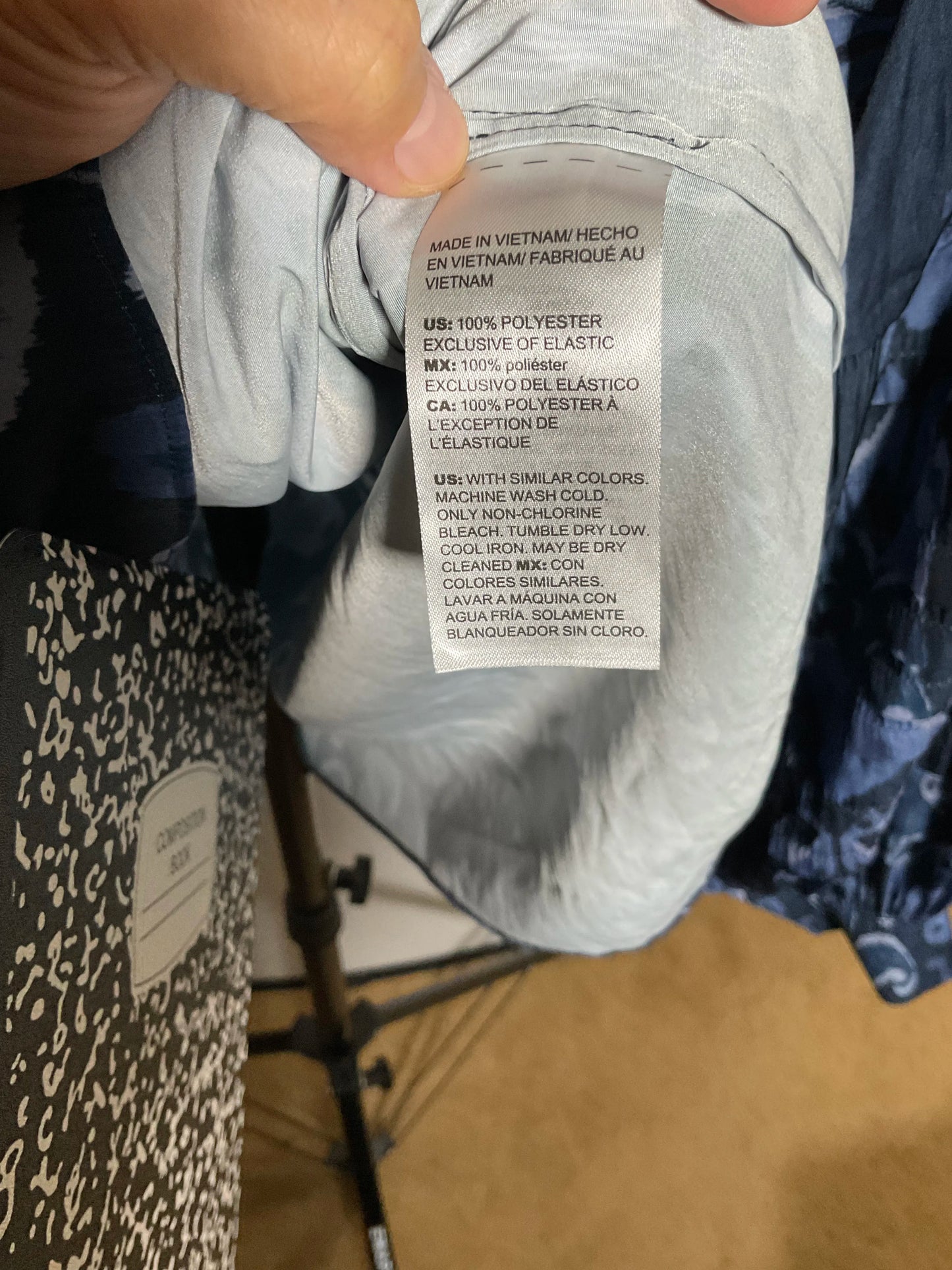A close-up of a hand holding the collar of a light gray garment to show the inner care label, with other clothing items and a store floor visible in the background.