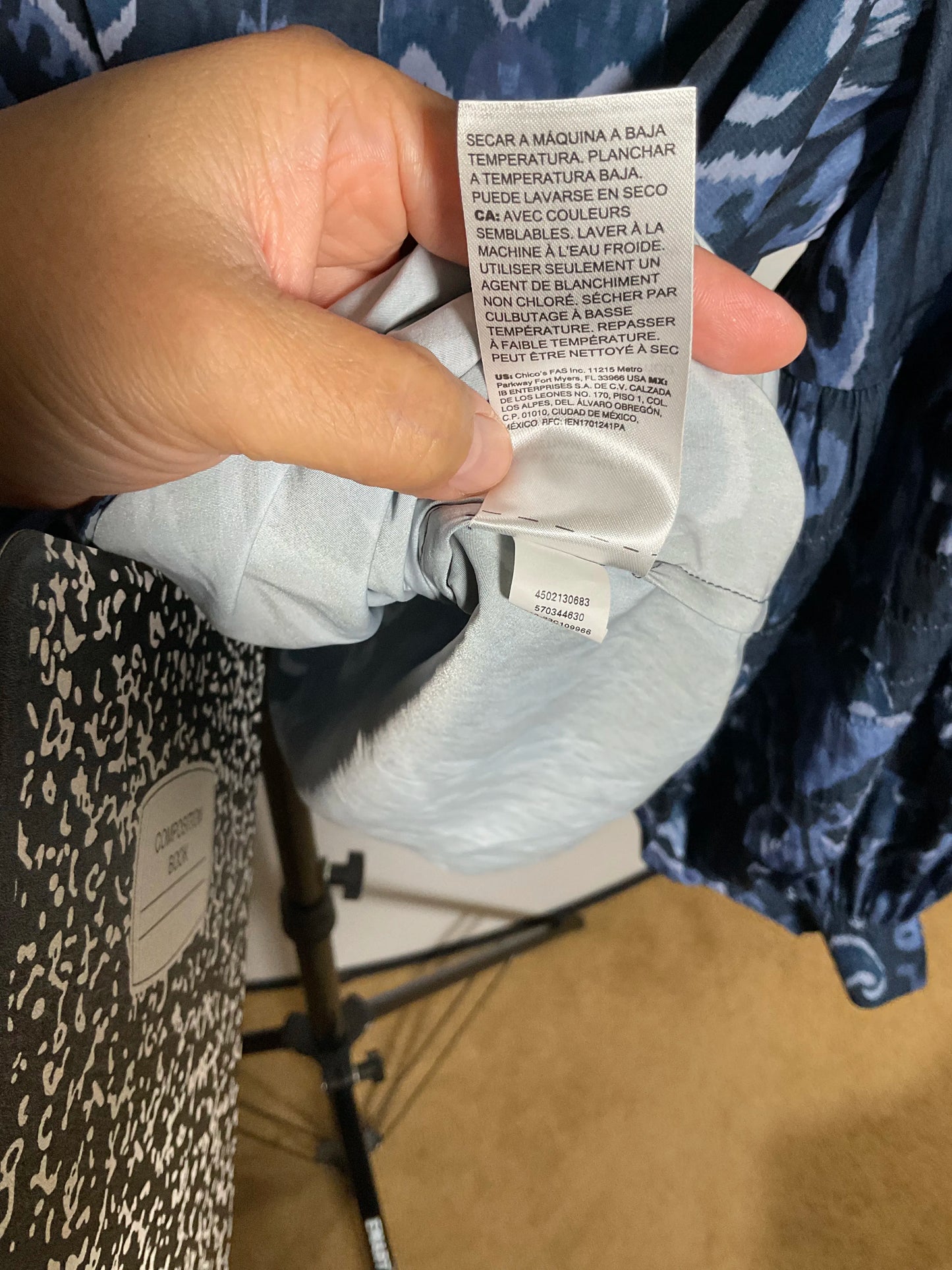 A close-up of a hand holding a white garment care label attached to a light blue-gray shirt, with patterned clothing and a tripod leg visible in the background.