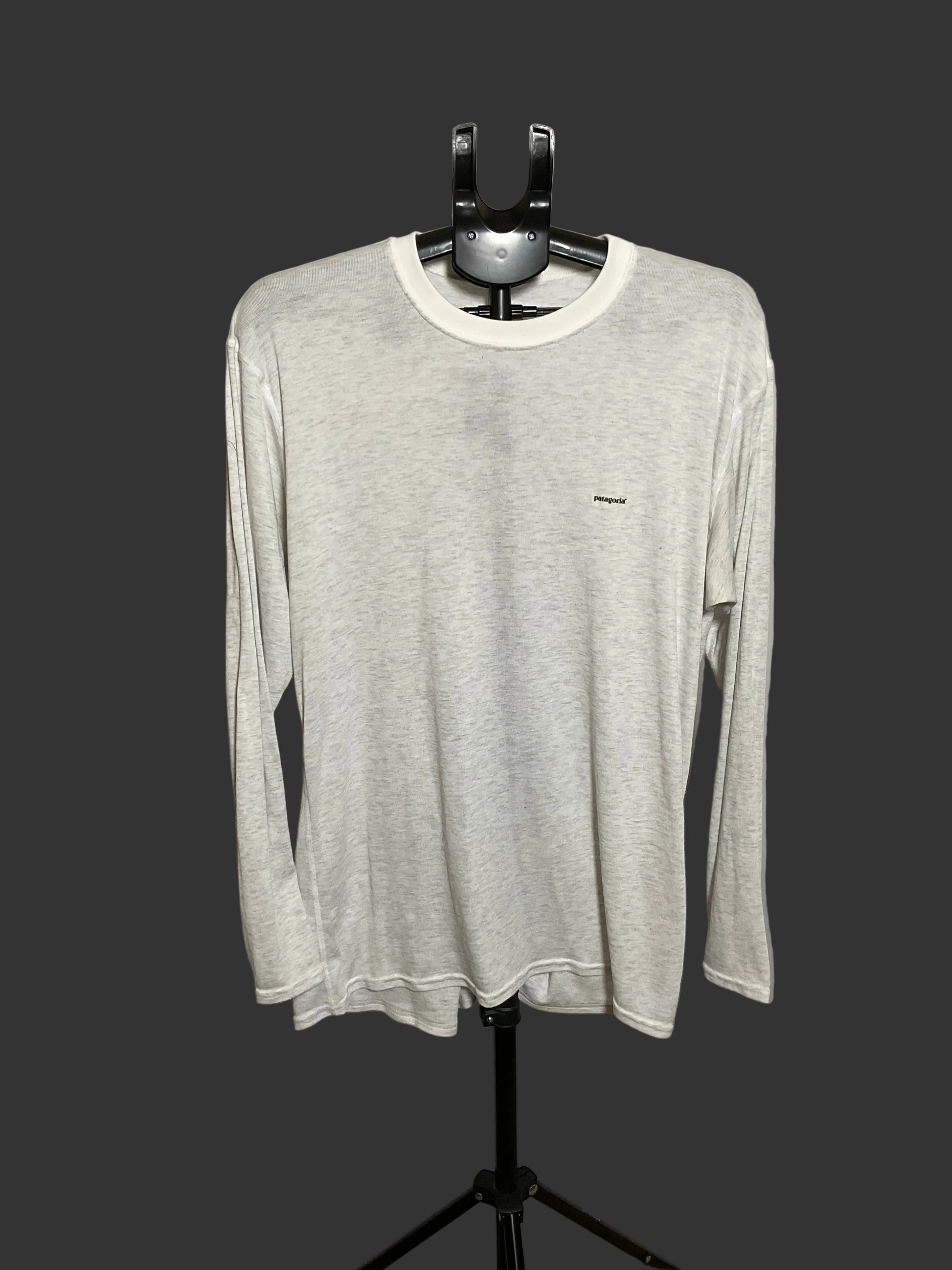 A light gray long-sleeve crewneck shirt with a small black logo on the left chest, displayed on a black hanger stand against a dark gray background.