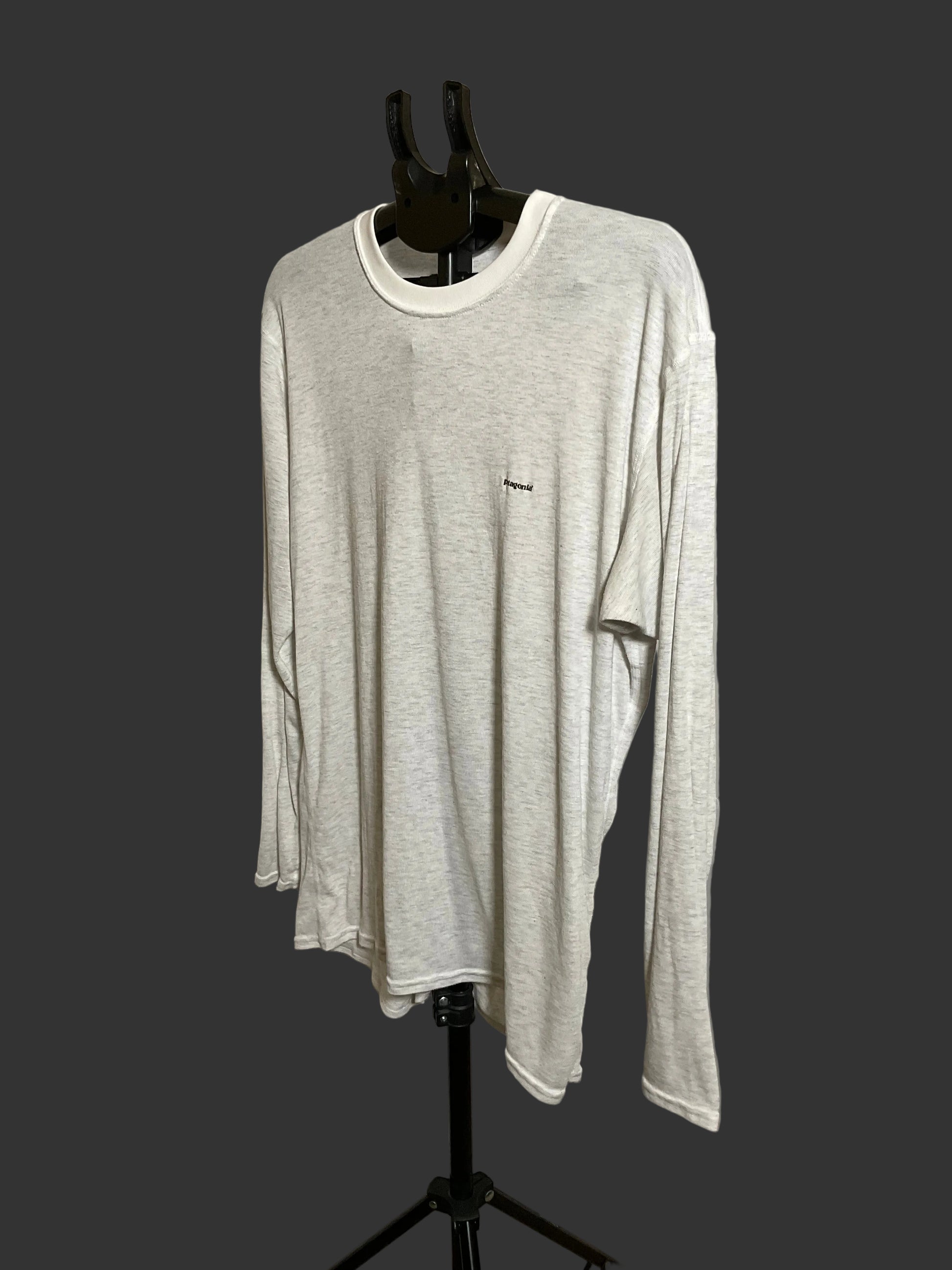 A light heather gray long-sleeve crewneck shirt with a small black embroidered logo on the left chest, displayed on a black hanger stand against a dark gray background.