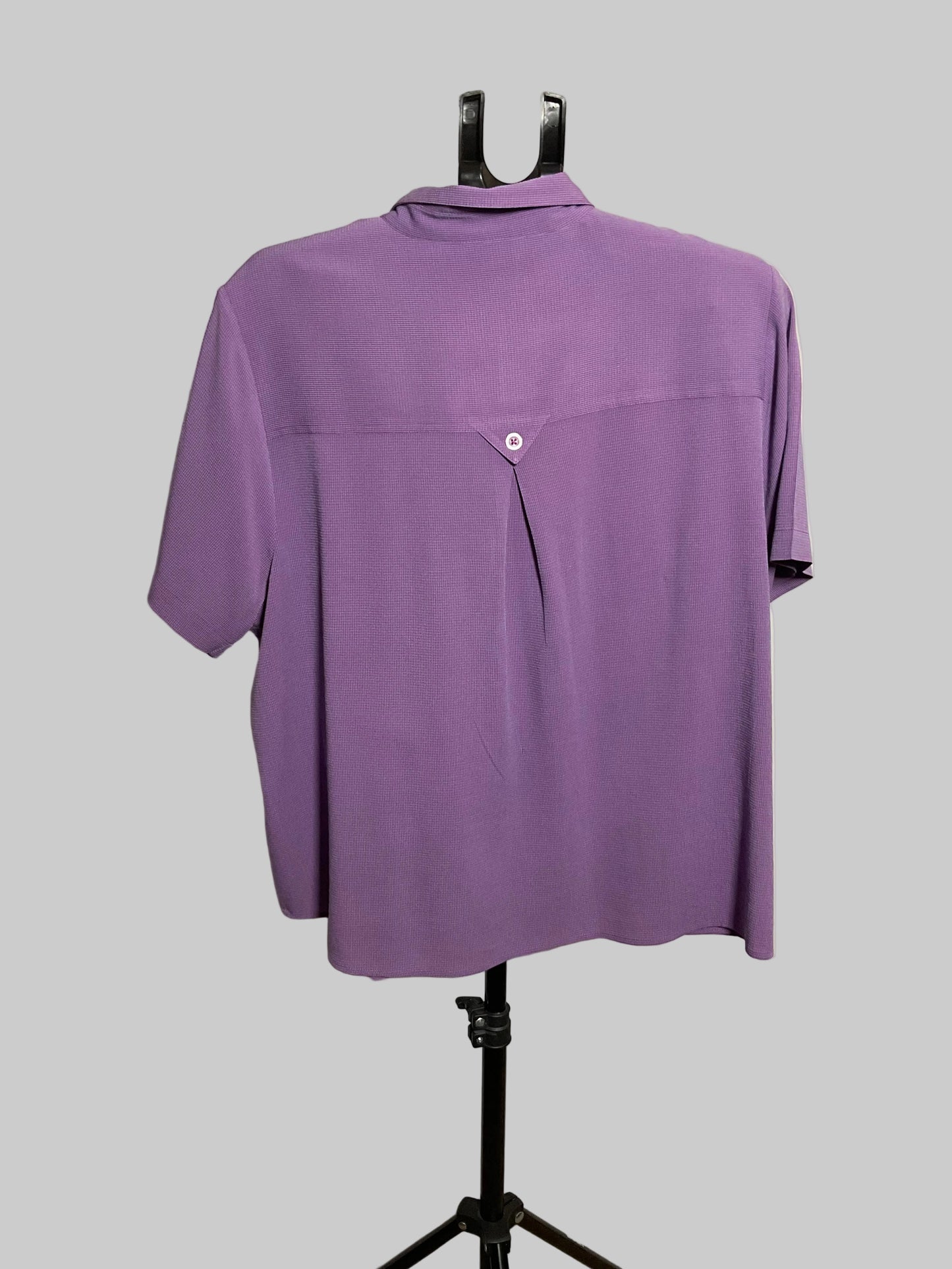 A purple short-sleeve button-up shirt displayed on a black dress form stand against a plain off-white wall.