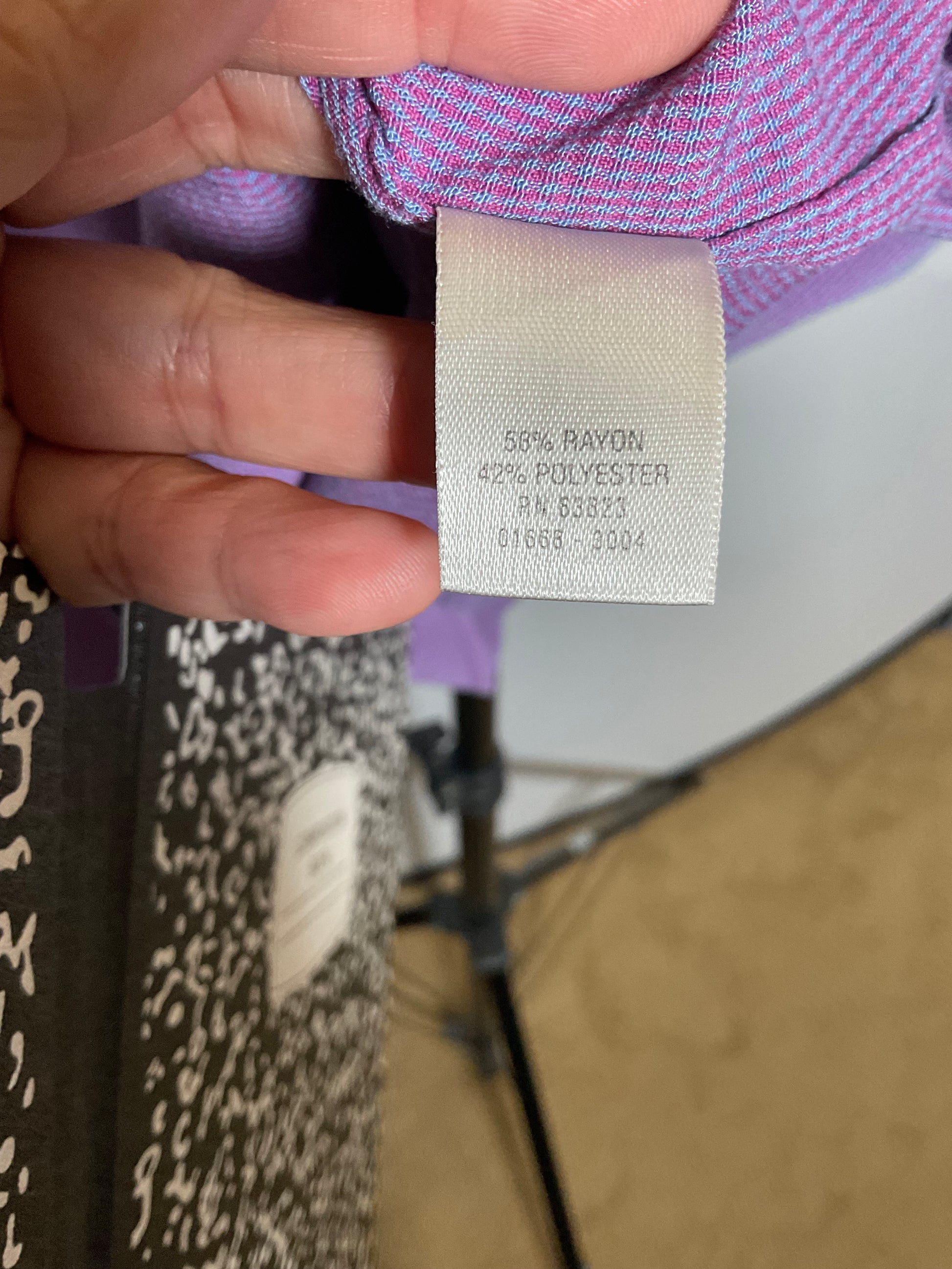 A close-up of a hand holding a fabric tag that reads '55% RAYON 45% POLYESTER' on a purple patterned garment, with a studio light stand visible in the background.