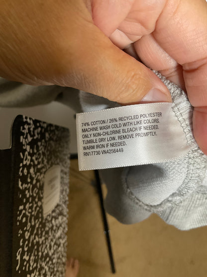 A close-up of a hand holding a white fabric care label sewn onto a light gray garment. The label contains printed care instructions and material composition text.