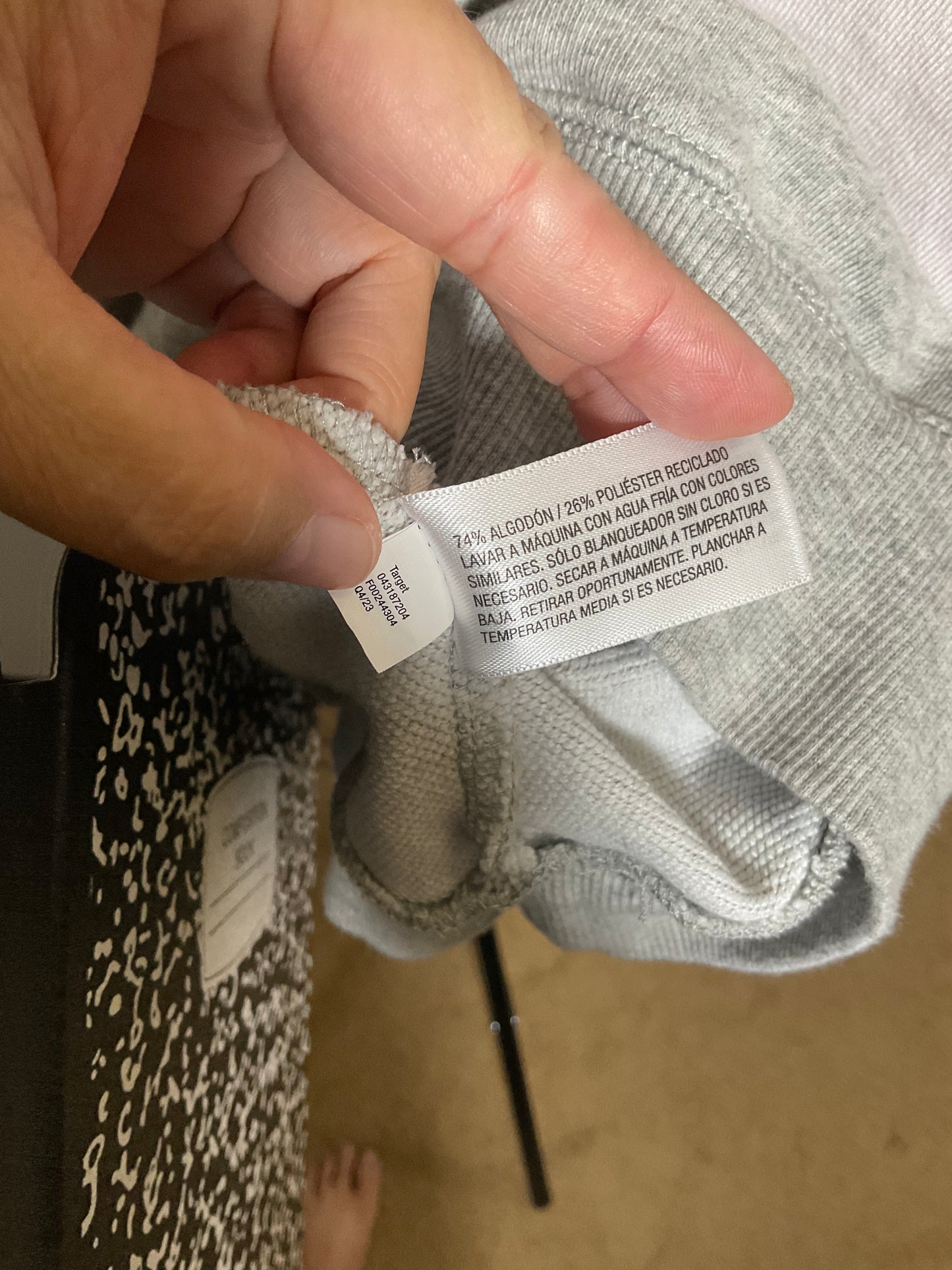 A close-up of a hand holding a white fabric care label attached to a light gray ribbed garment, with part of a patterned bag and a beige floor visible in the background.