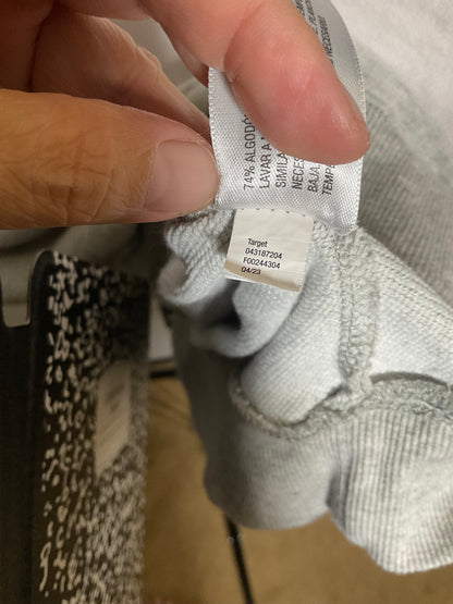 A close-up of a hand holding a white garment tag attached to a light gray ribbed knit garment, showing the brand name Target and size information.