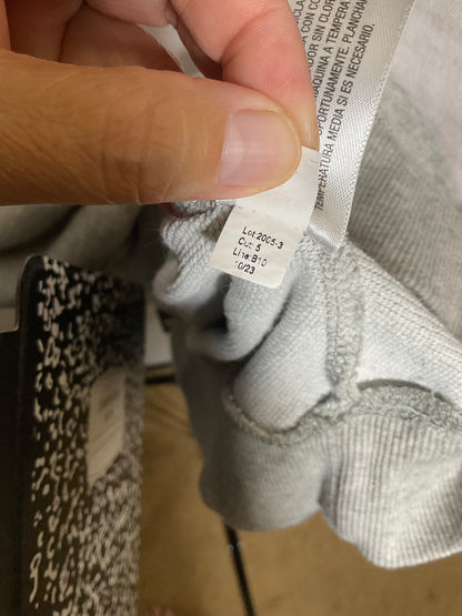 A close-up of a hand holding a white size tag attached to a light gray ribbed garment, with part of a black-and-white patterned item visible at the lower left.
