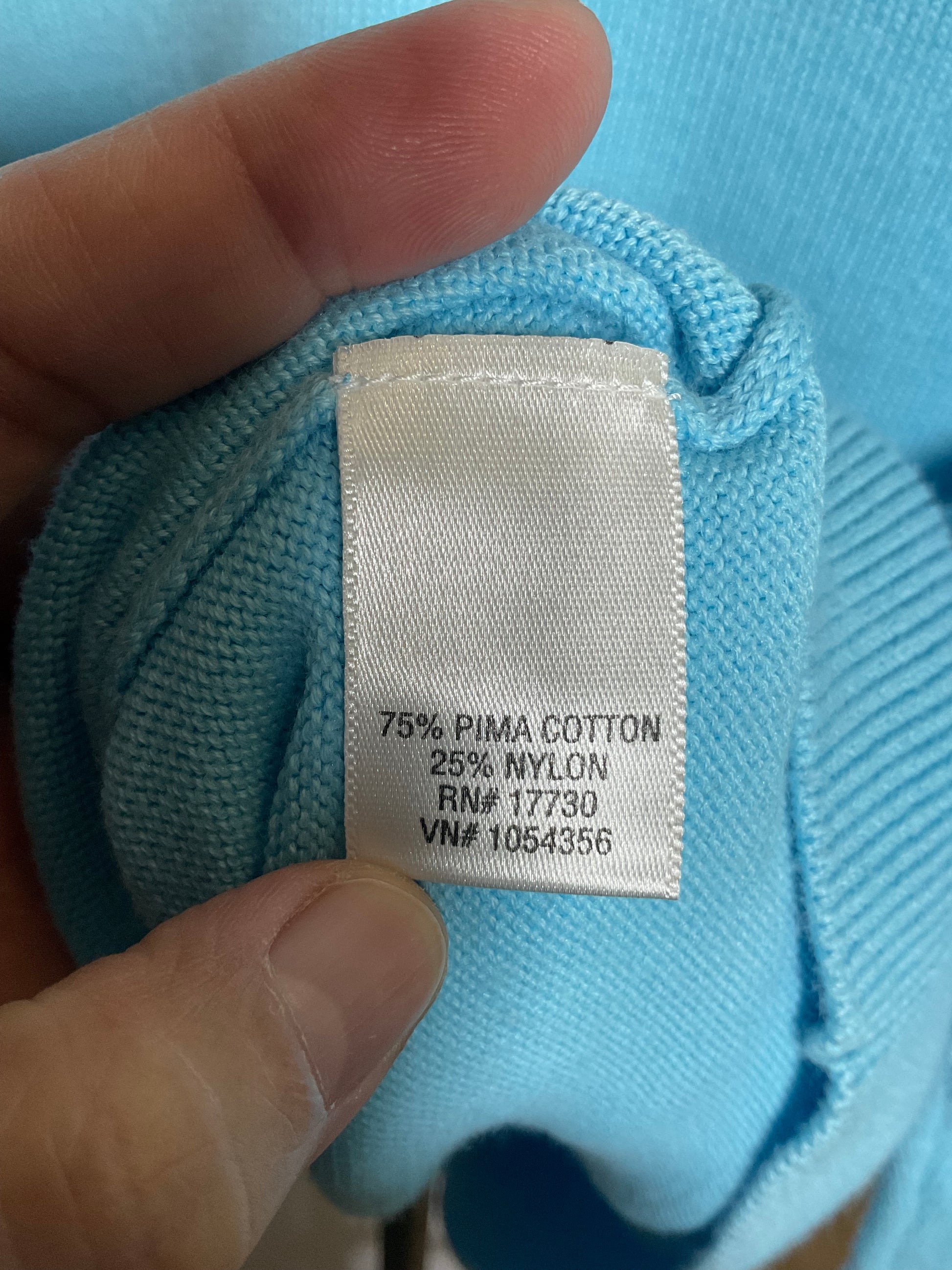 A close-up of a light blue knit garment held by a hand, showing a sewn-on fabric label with text reading '75% PIMA COTTON 25% NYLON RN# 17730 VN# 1054356'.