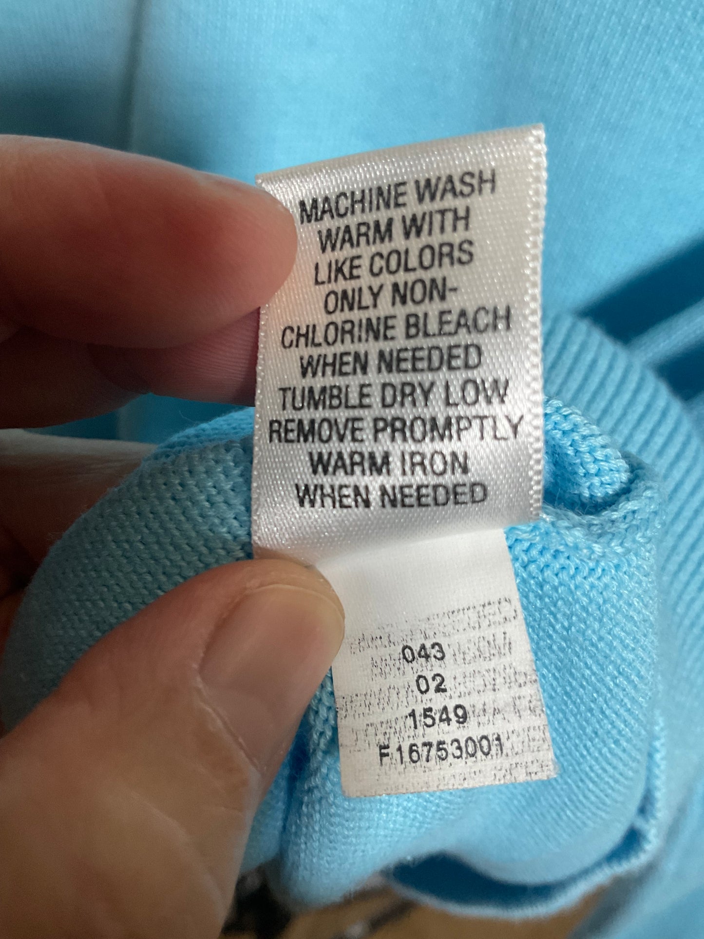 A close-up of a hand holding a white garment care label attached to a light blue knit fabric, showing washing instructions in black text.