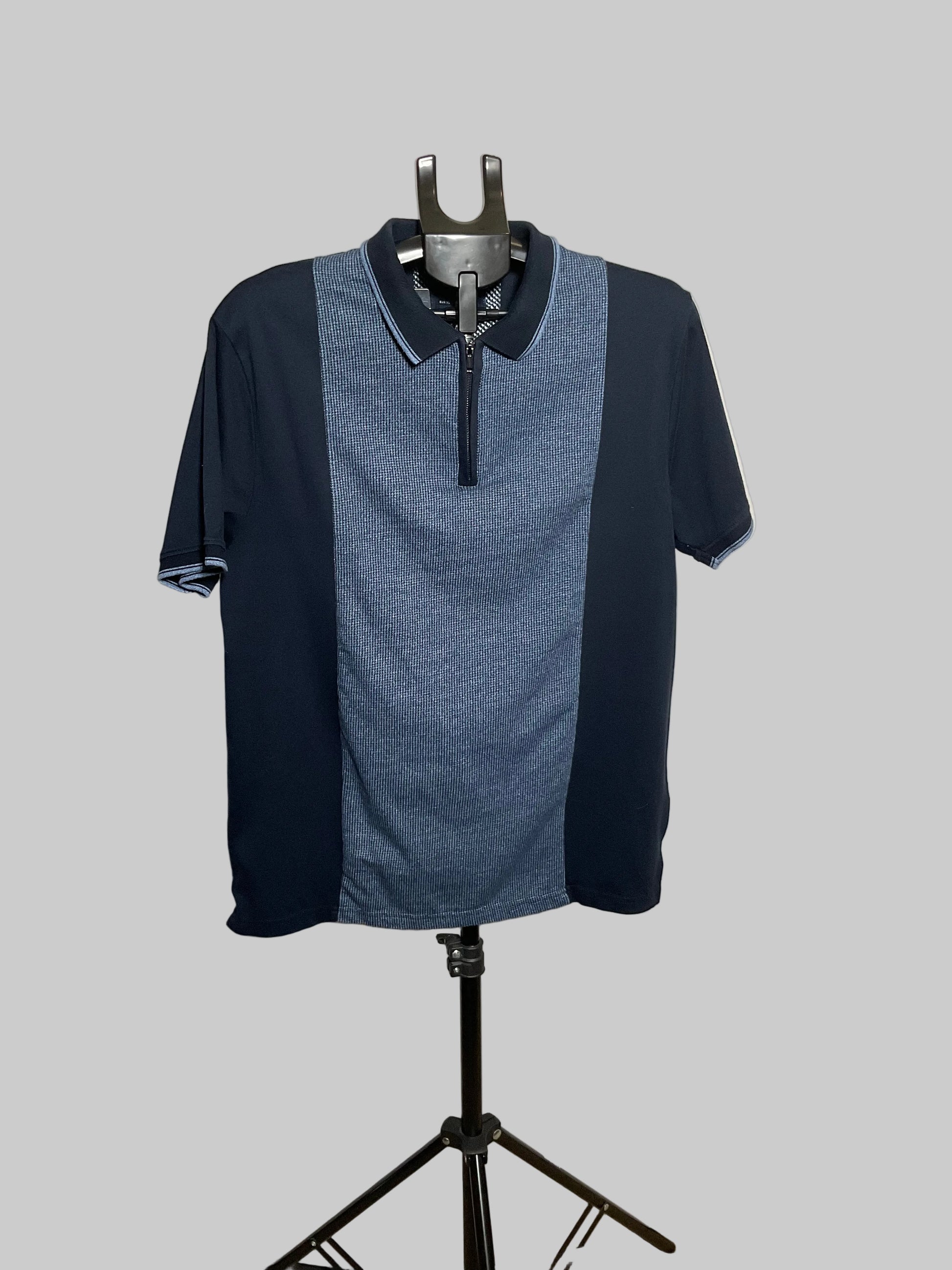 A men's short-sleeve polo shirt with a colorblock design, featuring a heathered blue center panel and solid dark navy side panels and collar, displayed on a black mannequin stand against a white studio backdrop.