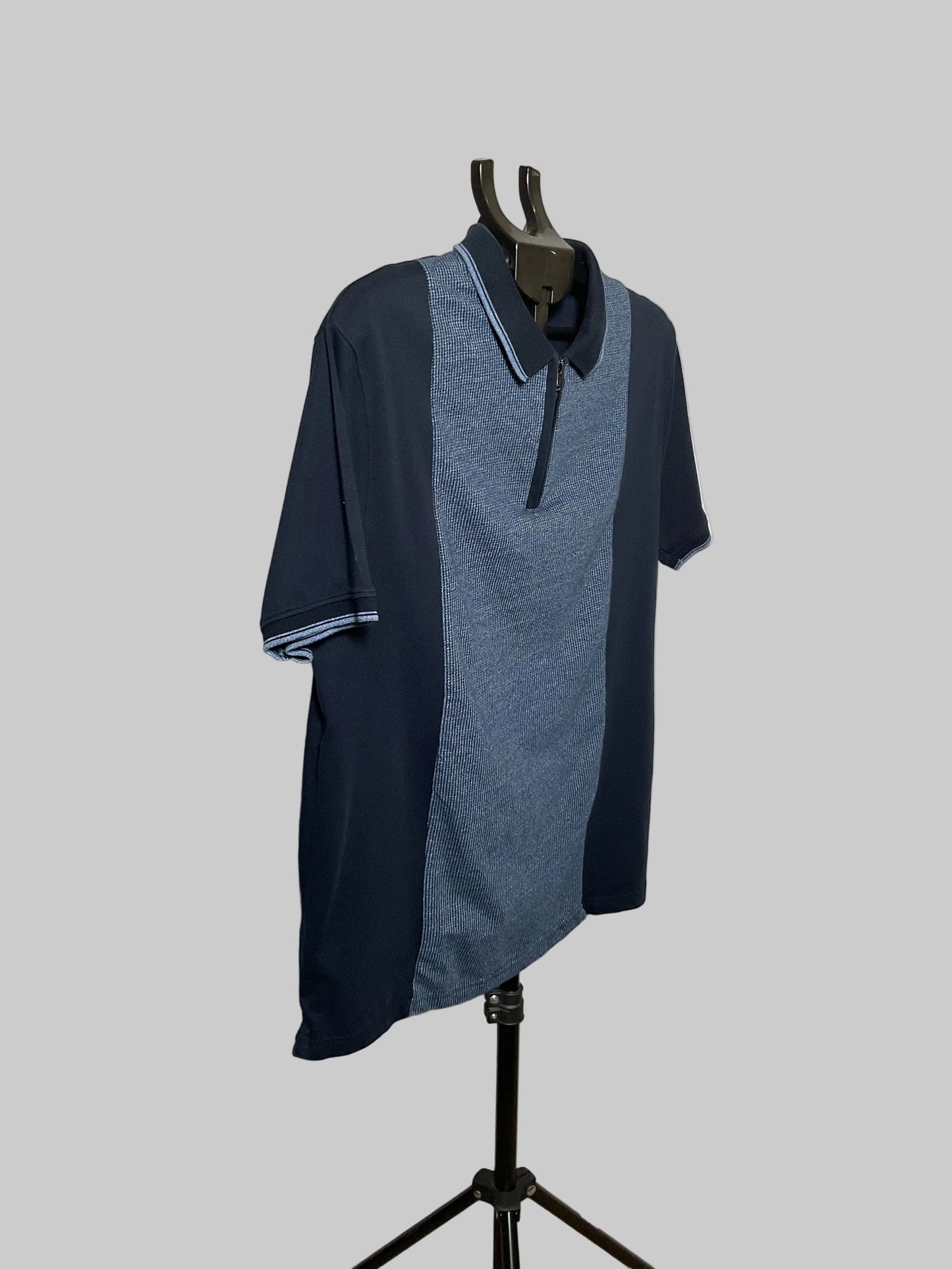 A short-sleeve polo shirt displayed on a black hanger stand against a white studio backdrop. The shirt has a color-blocked design with dark navy sleeves and side panels, a medium blue textured center panel, and light blue striped trim on the collar and sleeve cuffs.