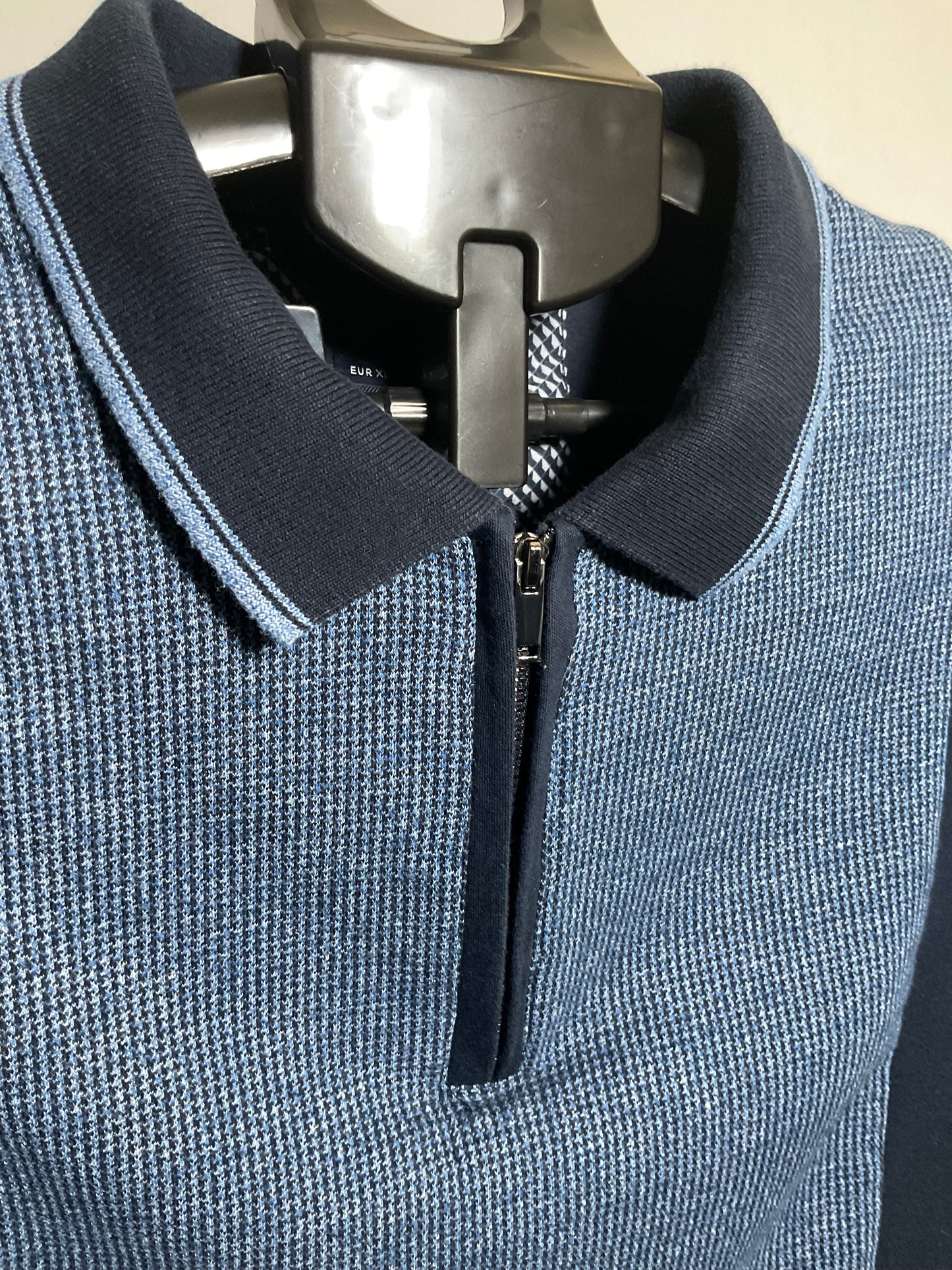 A close-up of a men's blue and navy polo shirt with a textured knit pattern, featuring a half-zip collar and a silver metal hanger.