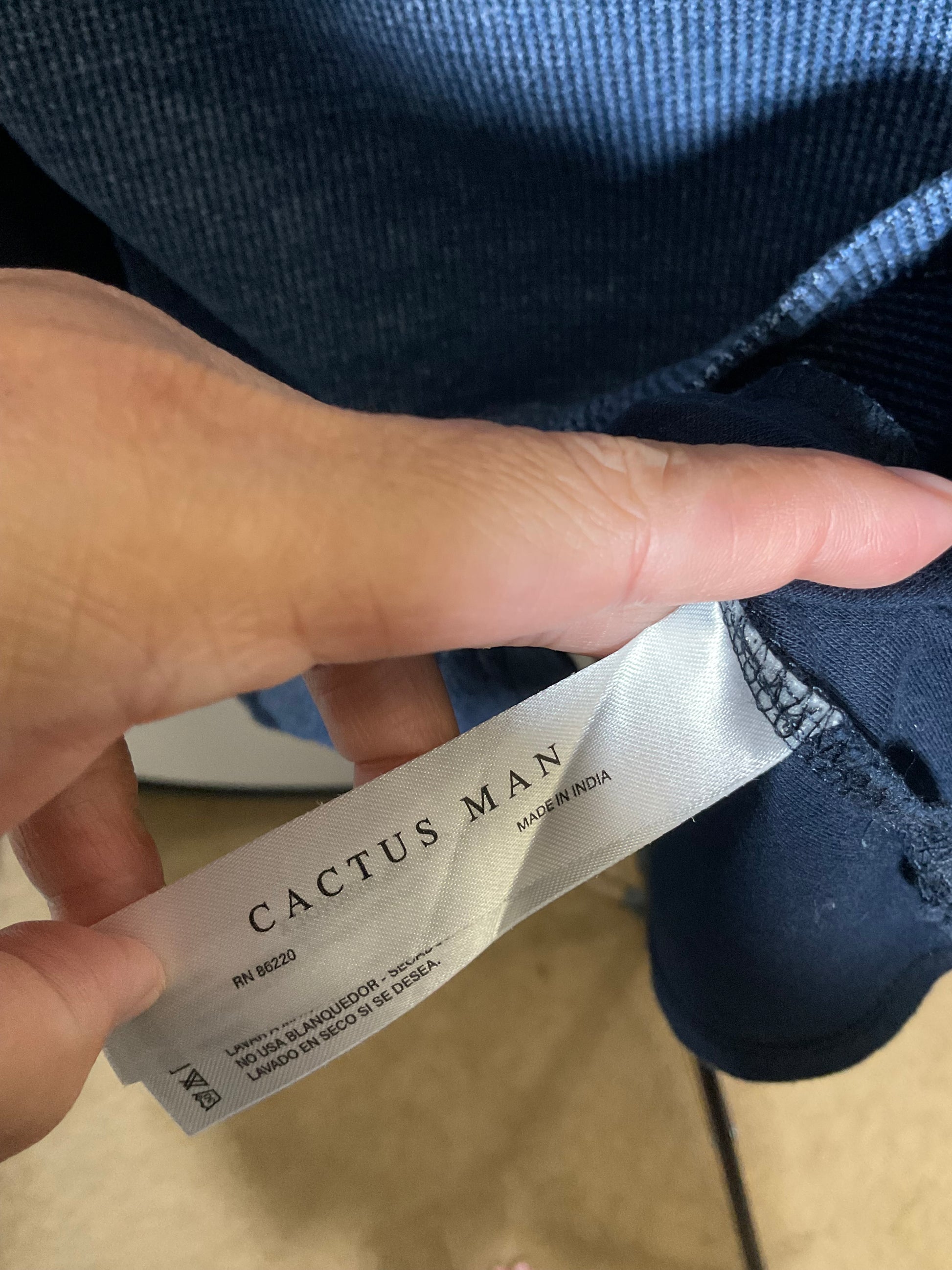 A close-up of a hand holding a white Cactus Man size tag next to the hem of a dark blue knit garment, showing the brand label and size information.