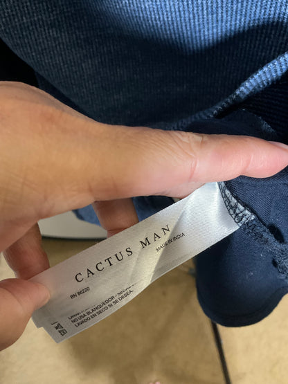 A close-up of a hand holding a white Cactus Man size tag next to the hem of a dark blue knit garment, showing the brand label and size information.
