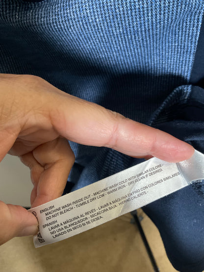 A close-up of a hand holding a white fabric care label attached to a blue ribbed garment, showing printed care instructions in English, Spanish, and French.