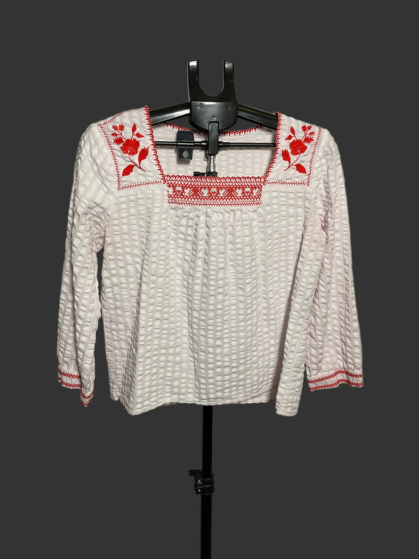 A white textured long-sleeve blouse with red floral and geometric embroidery on the neckline and cuffs, displayed on a black hanger stand against a plain gray wall.