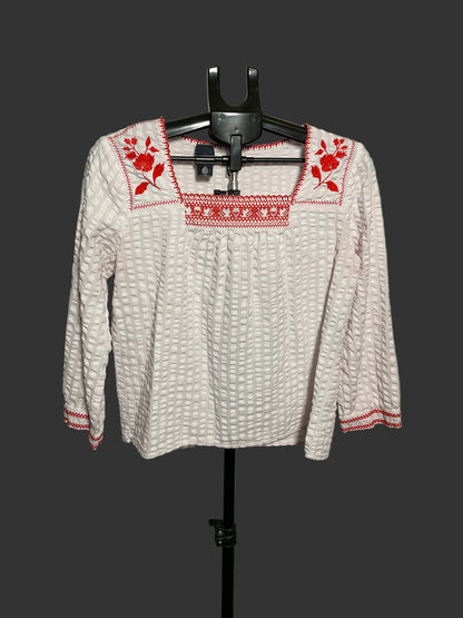 A white textured long-sleeve blouse with red floral and geometric embroidery on the neckline and cuffs, displayed on a black hanger stand against a plain gray wall.