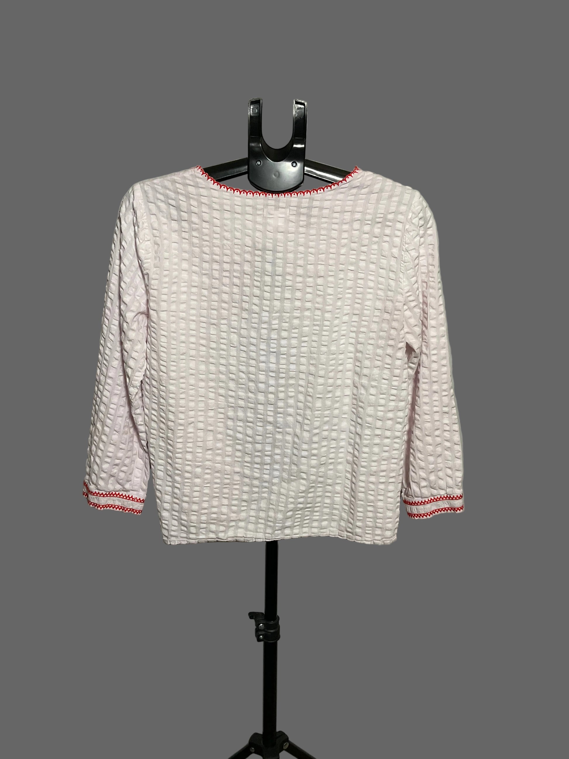 A light pink textured long-sleeve top with red trim on the collar and cuffs, displayed on a black hanger stand against a plain light gray background.