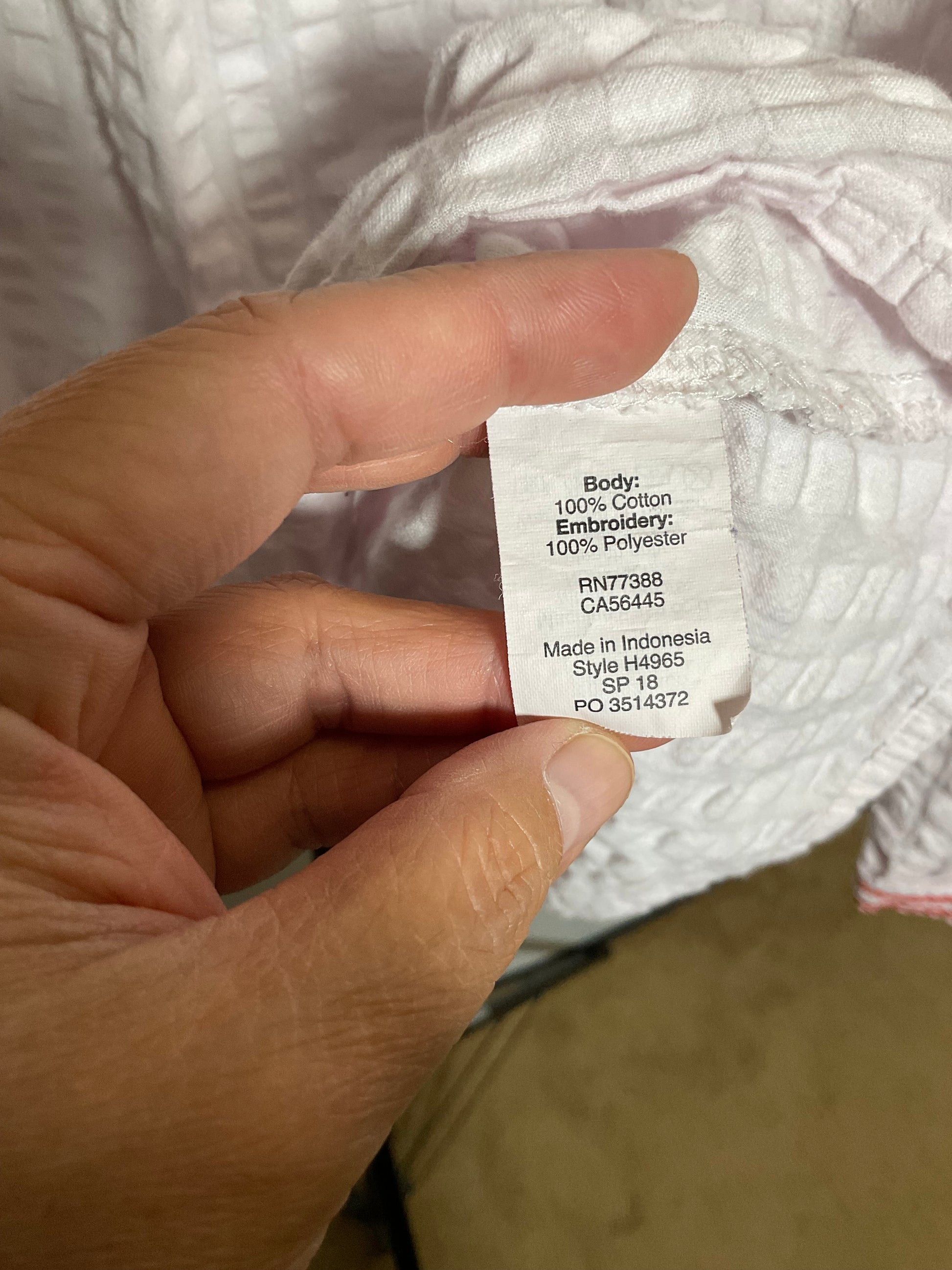 A close-up of a hand holding a white fabric care label that reads 'Body: 100% Cotton Embroidery: 100% Polyester Made in Indonesia Sija H4965' on a white textured garment.