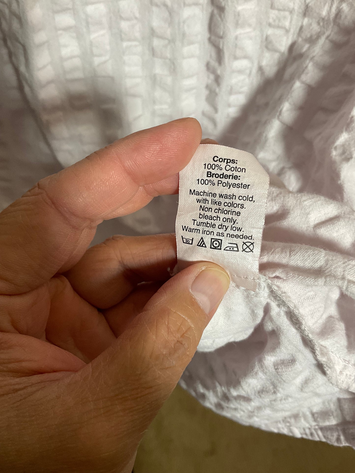 A close-up of a hand holding a white fabric care label with printed instructions, set against a textured white garment.