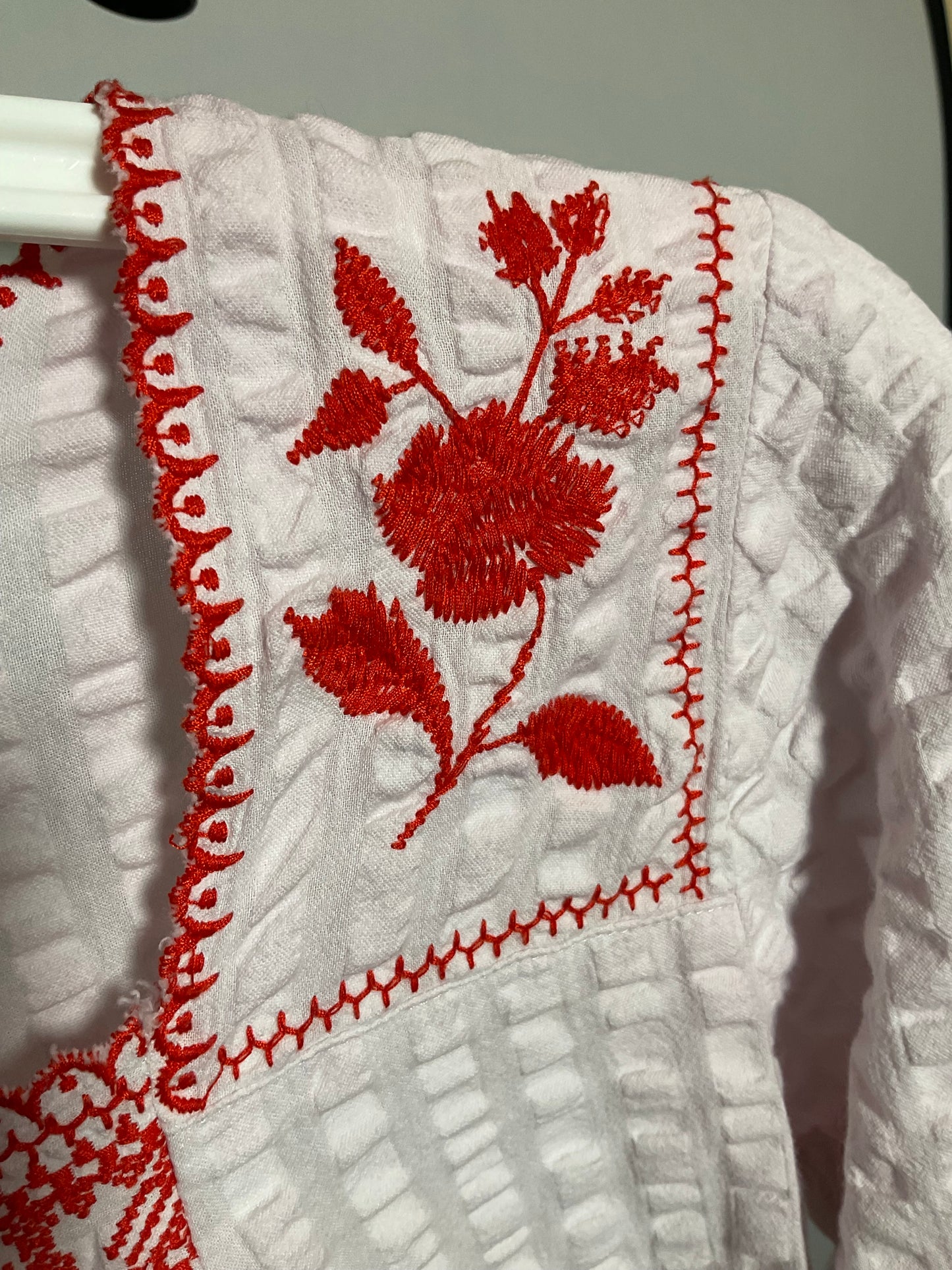 A close-up of a white textured garment featuring vibrant red floral embroidery and decorative red stitched borders.